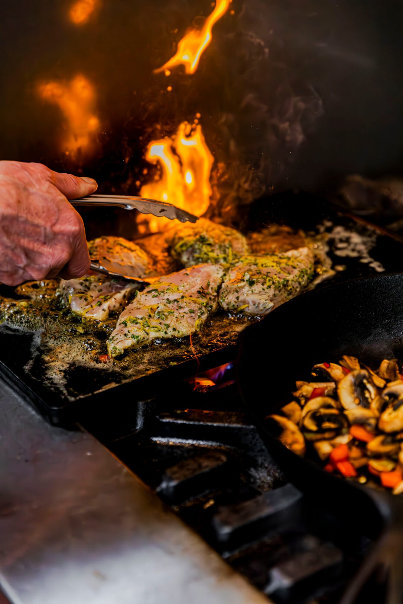 Person's hand holding tongs cooking fish fillets over a flame, with vegetables in a pan nearby.