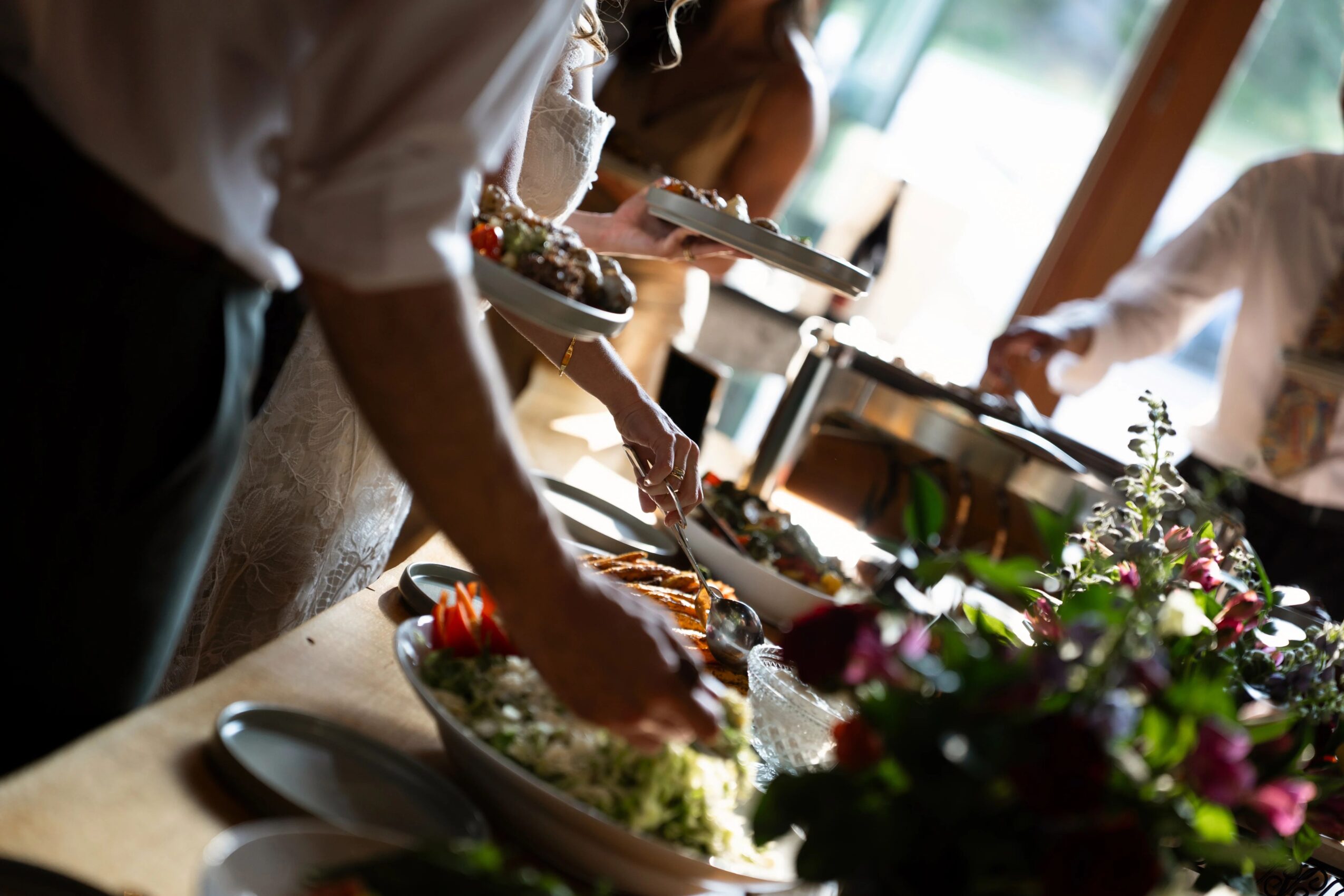 People serving food at a buffet table with flowers and various dishes, in a well-lit room.