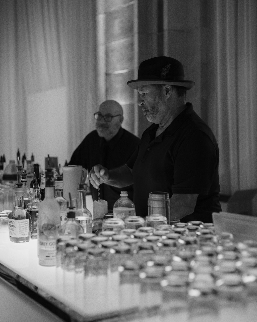 Two men behind a bar with bottles and jars, one wearing a hat, in a black-and-white setting.