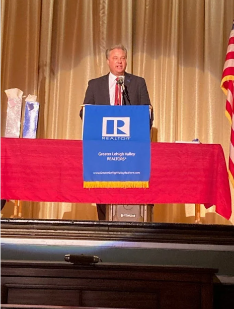 Man in suit speaking at a podium on stage with a gold curtain background and American flag to the right.