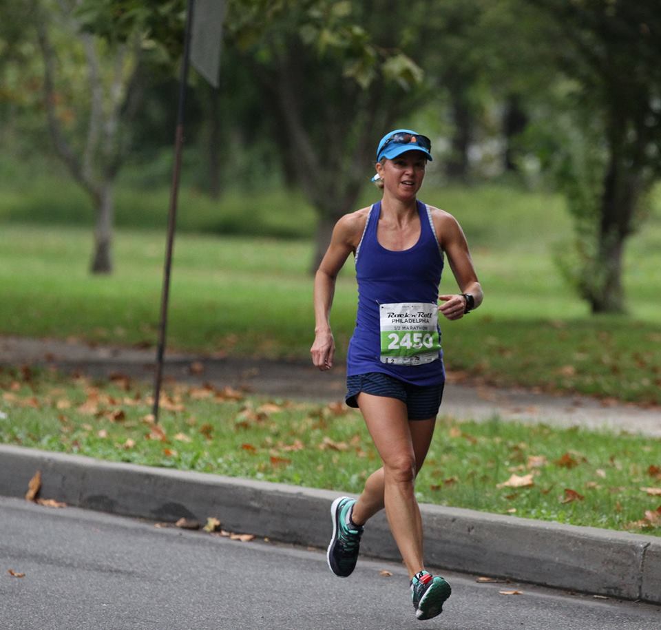 Woman running outdoors in a park, wearing a blue tank top, shorts, and a cap, with trees and grass in the background.