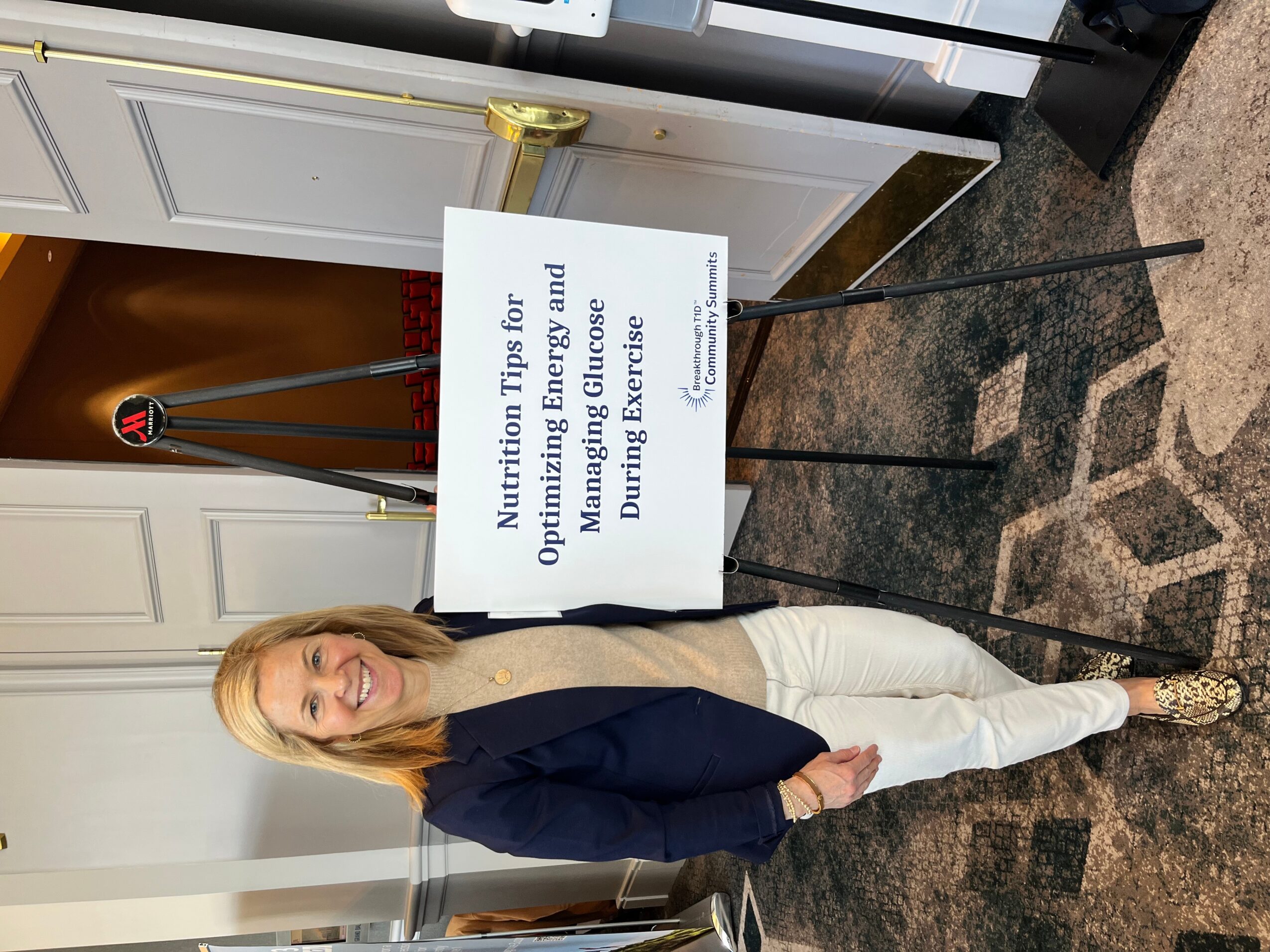 Woman standing next to a sign about nutrition tips for energy and performance, indoors with white cabinets and a patterned carpet.