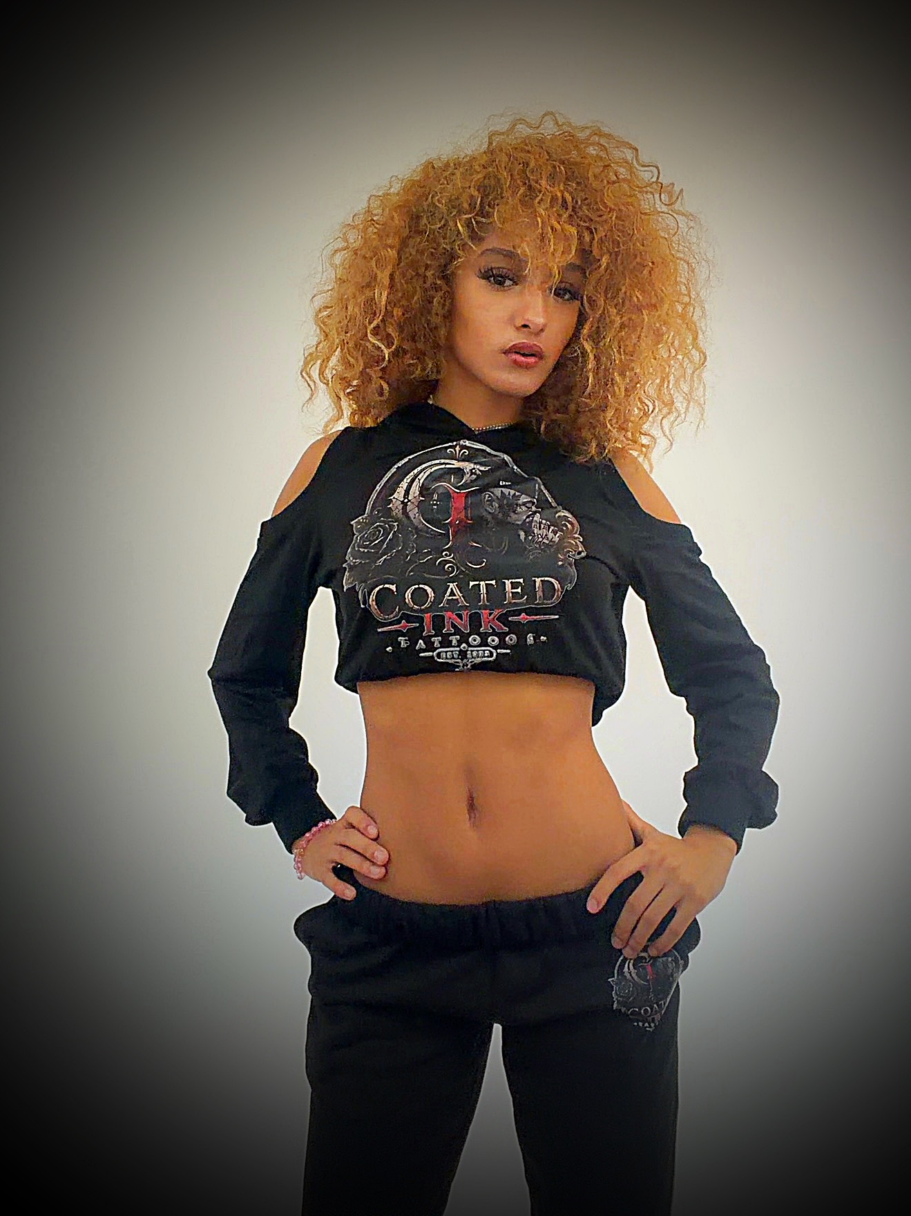 Woman with curly hair wearing a black crop top with graphic design and cold shoulder sleeves, standing with hands on hips.