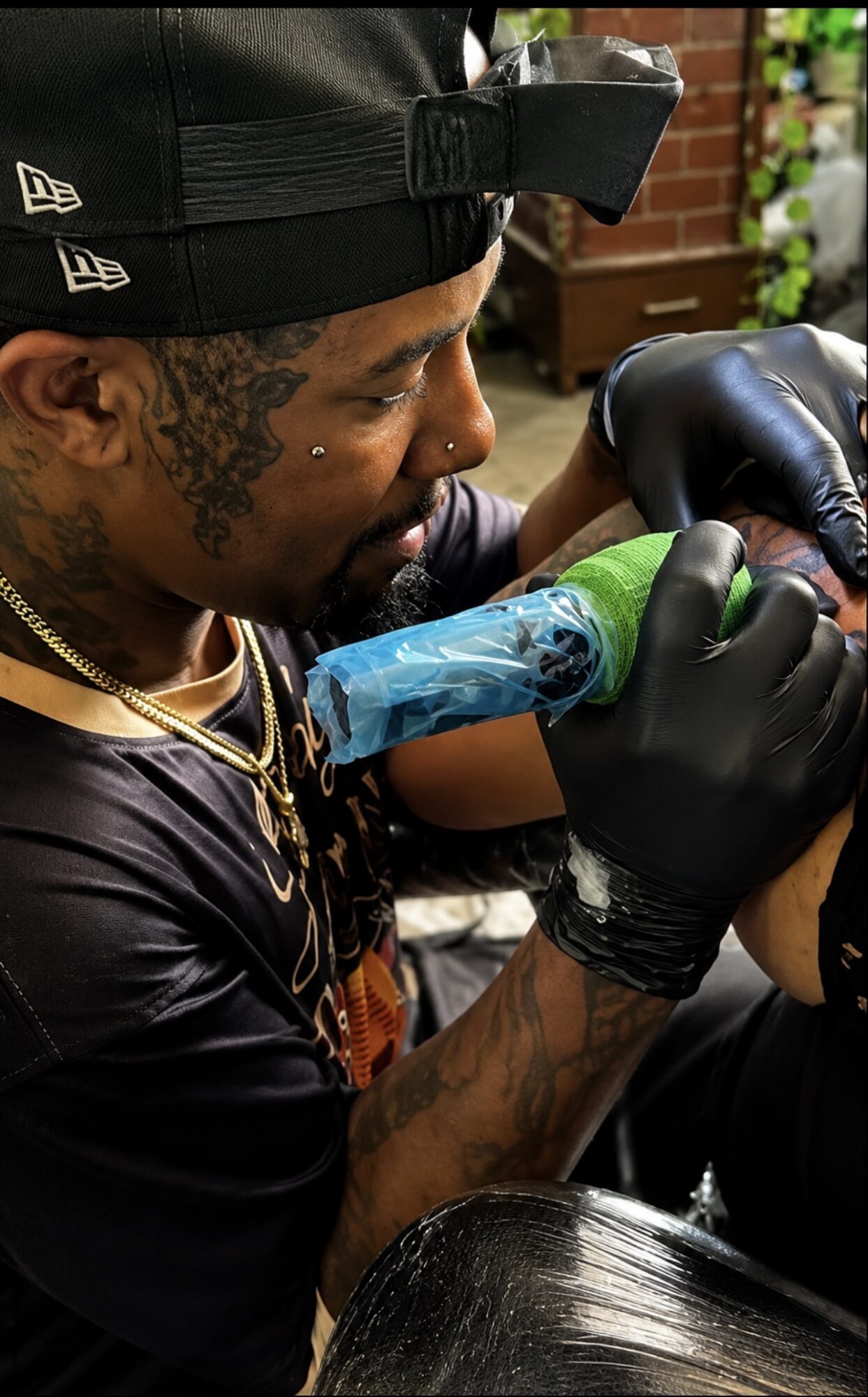 Tattooed man with a headlamp tattooing a person's arm in a tattoo studio, wearing black gloves and a black shirt.