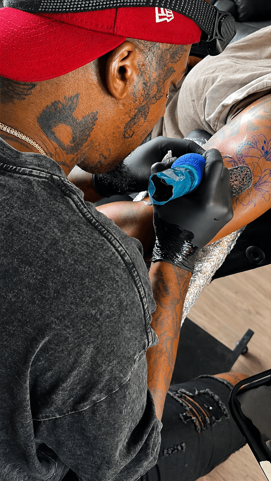 Tattoo artist in red cap tattooing a colorful design on a person's arm, wearing black gloves.