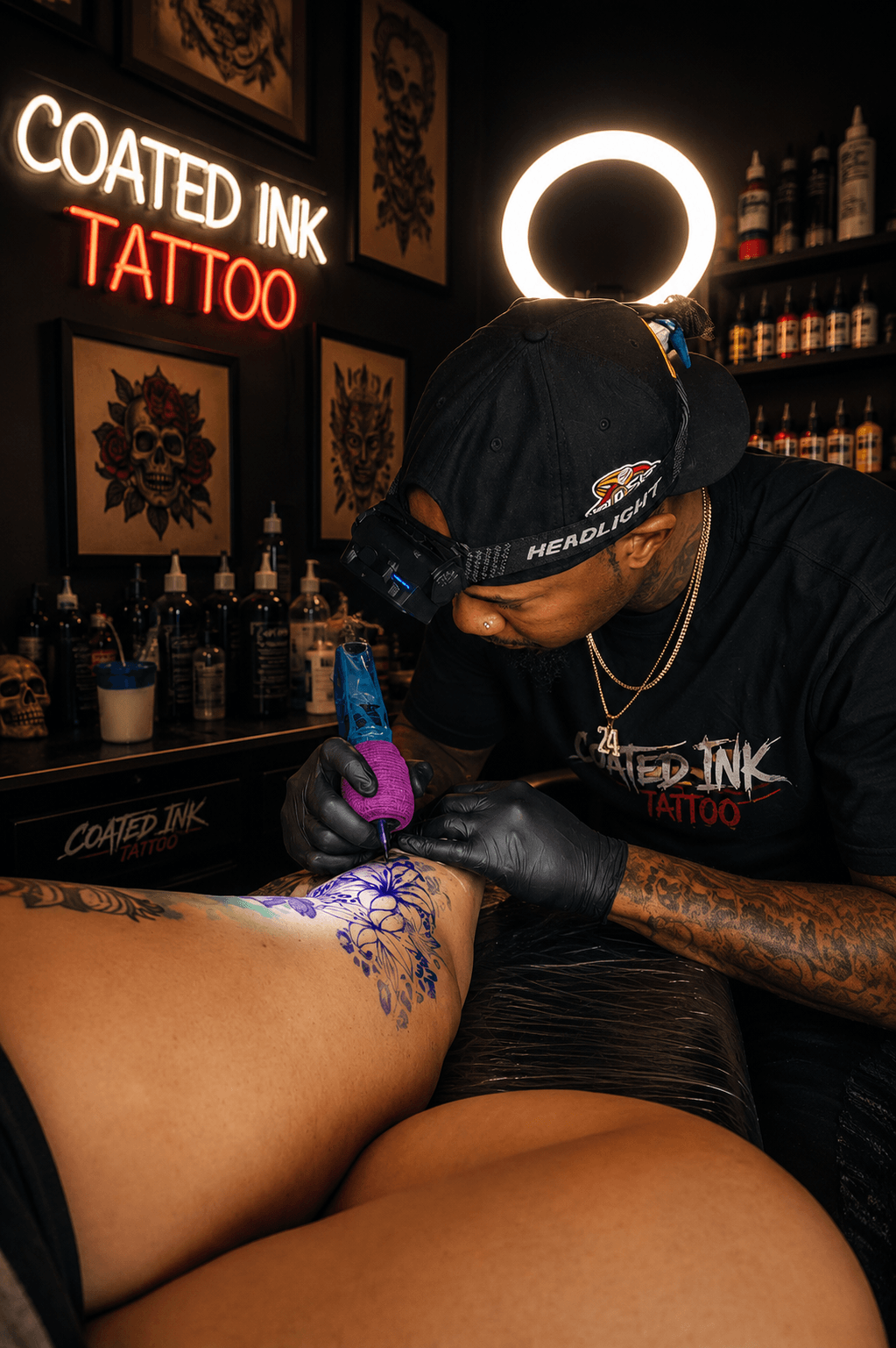Tattoo artist working on a person's leg in a tattoo studio with neon signs and shelves of ink bottles.