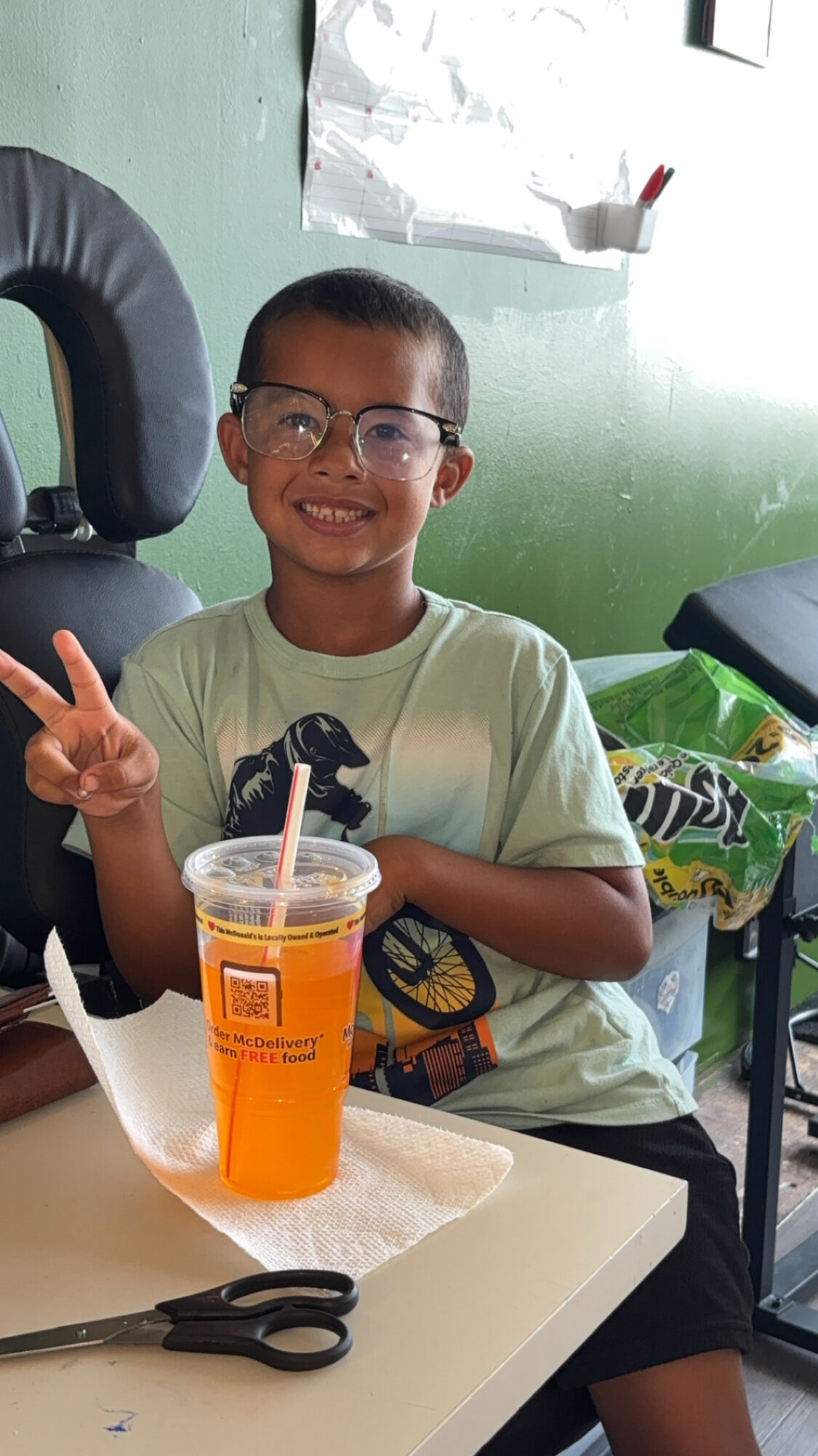 Child smiling and making a peace sign with a drink on the table, in a room with green walls.
