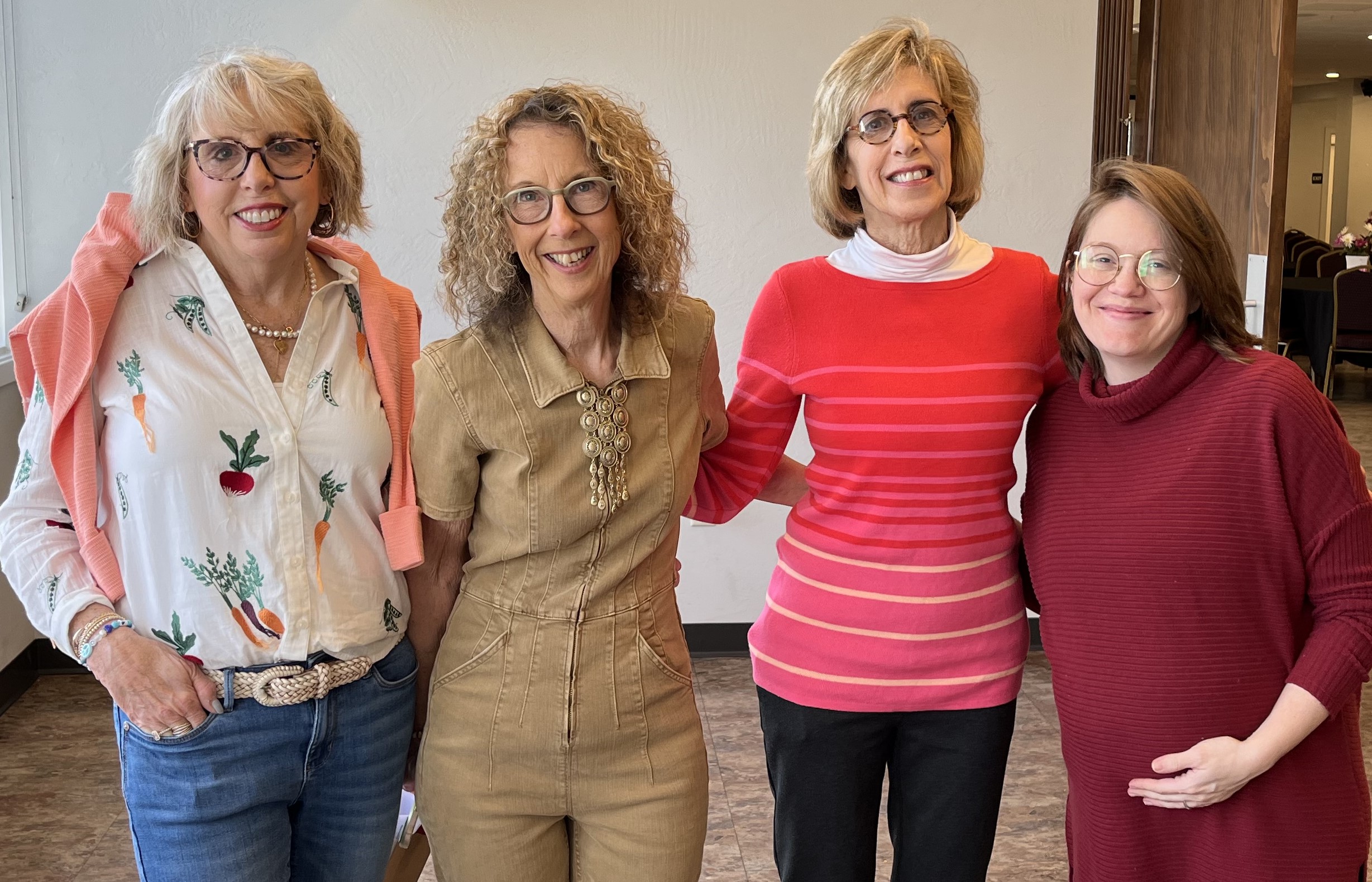 Four women standing together indoors, smiling, with a wooden door in the background.