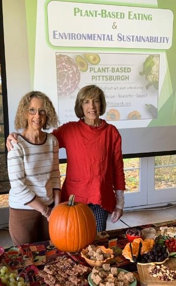 Two women stand behind a table with a pumpkin and food, in front of a presentation screen about plant-based eating and sustainability.