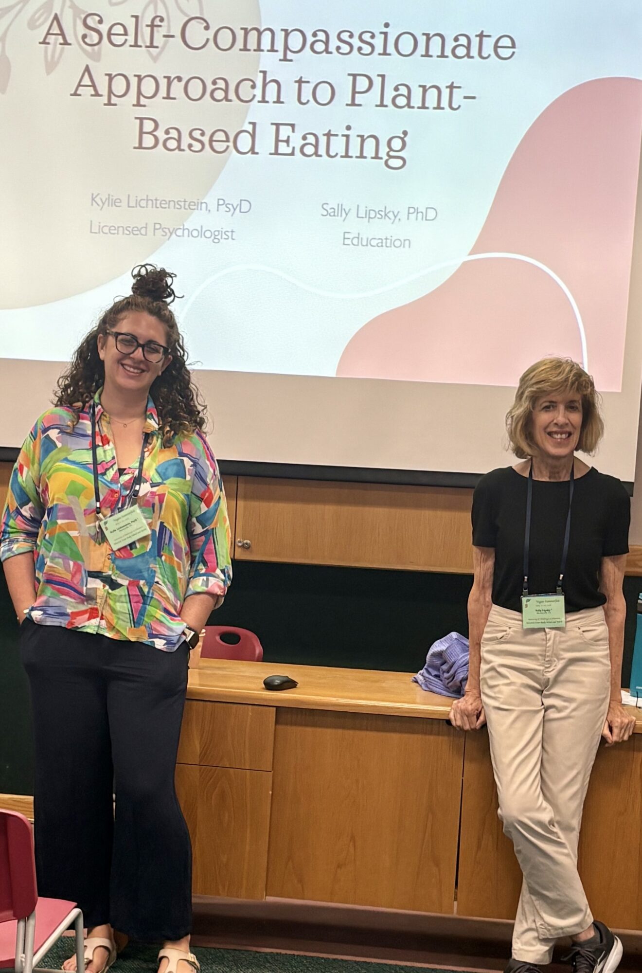 Two women standing in front of a presentation screen, smiling, in a classroom setting.
