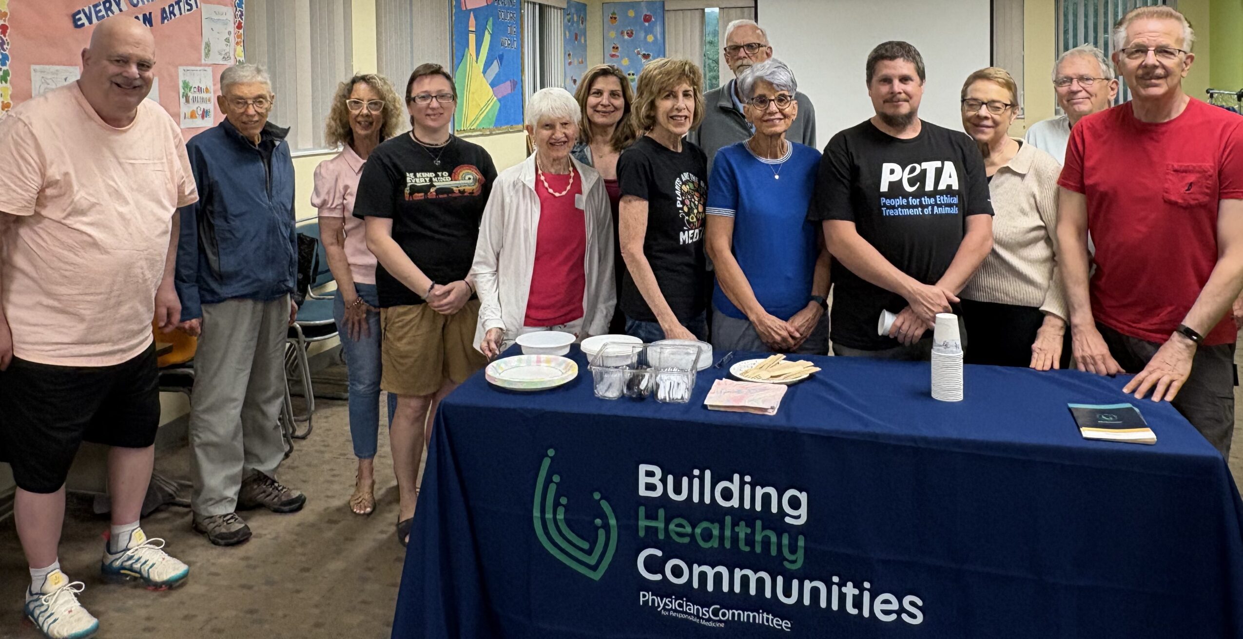 Group of people standing behind a table with a banner that reads 'Building Healthy Communities' in a room with windows and curtains.