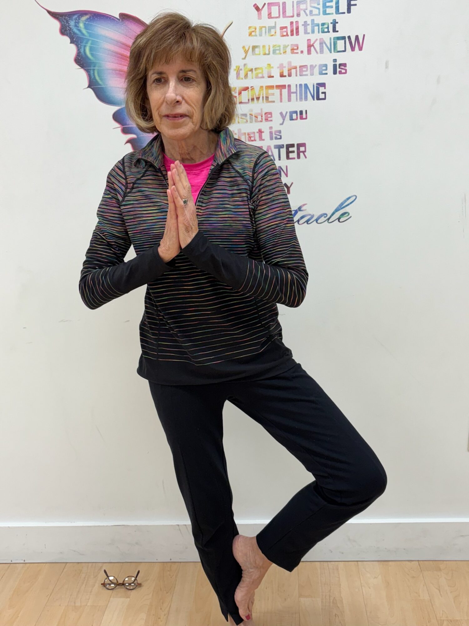 Woman in yoga pose with hands together, standing on one leg, in front of a wall with colorful text and a butterfly illustration.