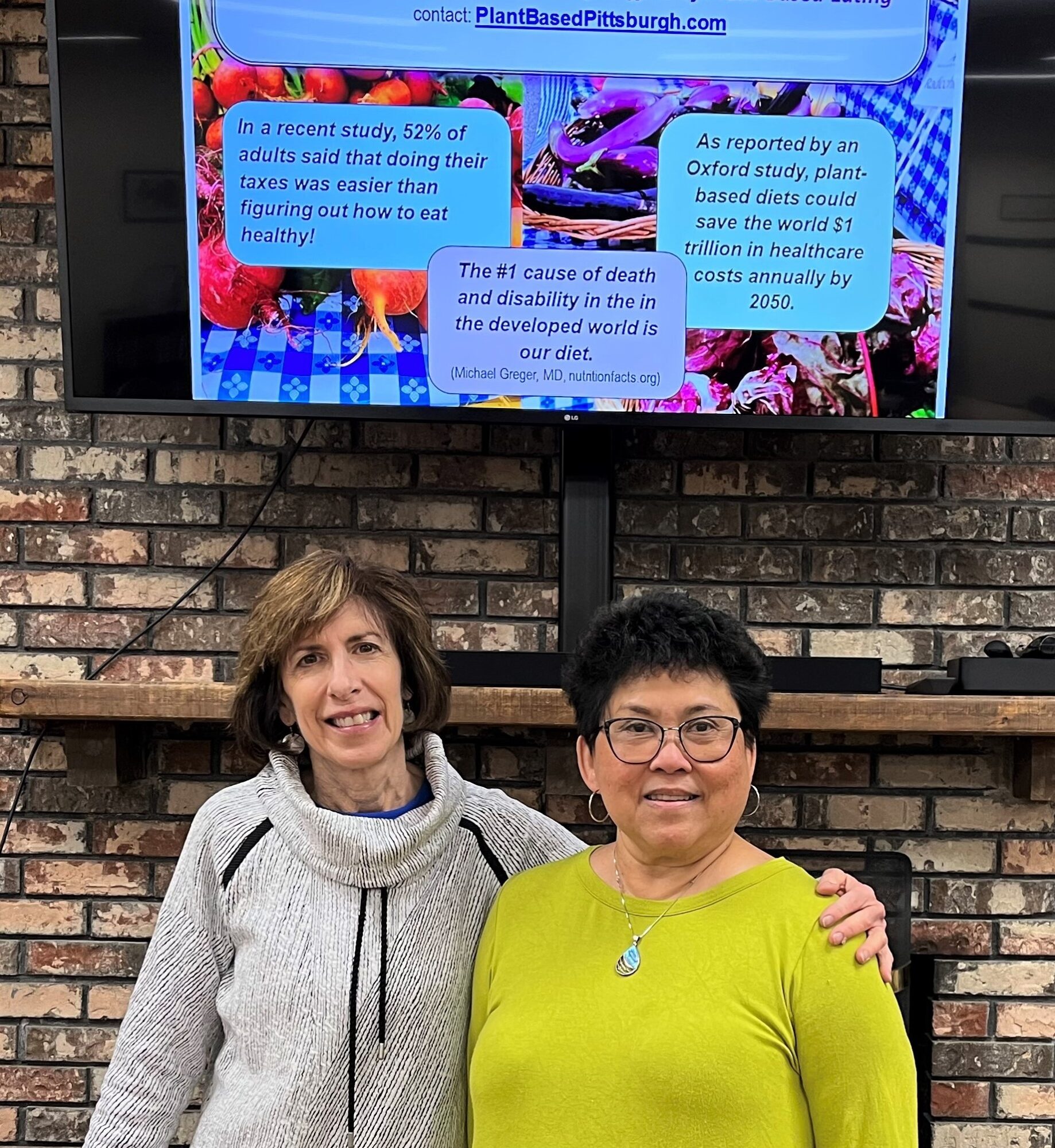 Two women standing in front of a brick wall, smiling, with a screen displaying text behind them.