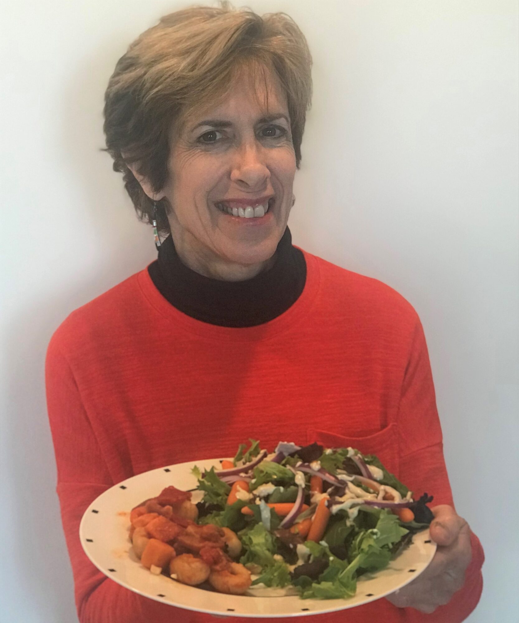 Woman with short hair smiling, wearing a red top with a black collar, holding a plate of salad.