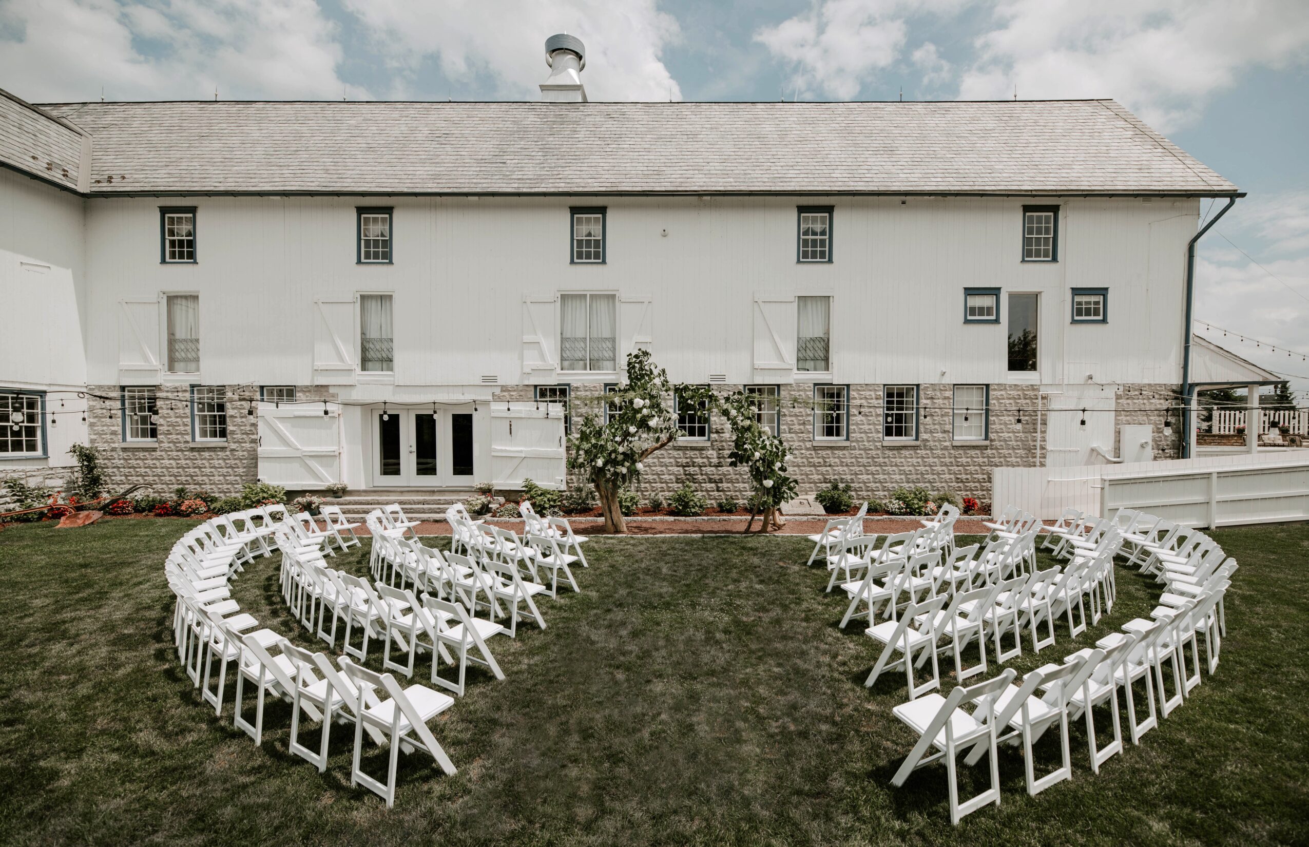 White chairs arranged in a semi-circle on grass in front of a large white building with small windows and a stone foundation.