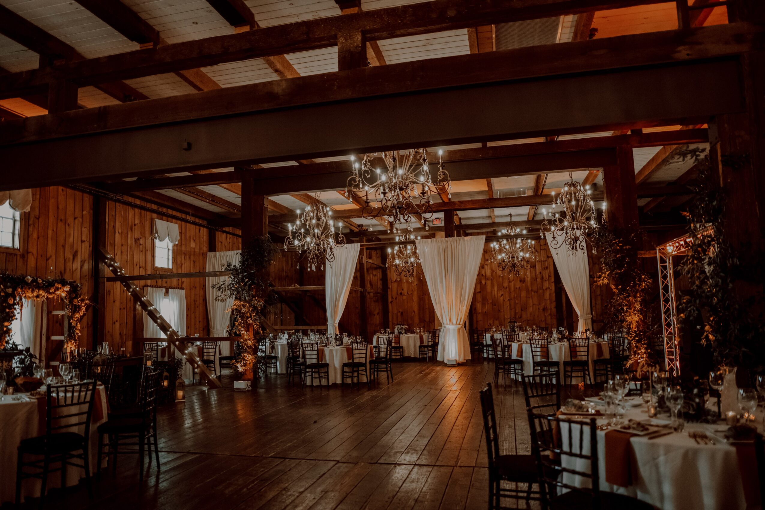 Elegant event space with chandeliers, draped curtains, and round tables with chairs, wooden floors, and warm lighting.