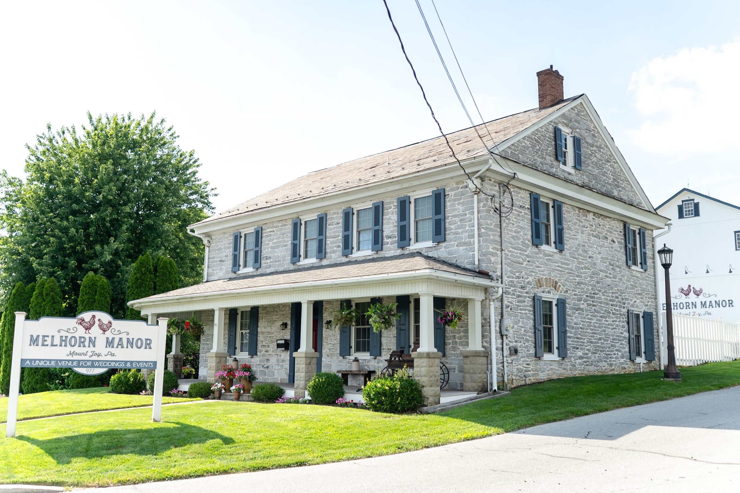 Stone house with front porch, blue shutters, and a sign reading Melhorn Manor, surrounded by a lawn and trees.