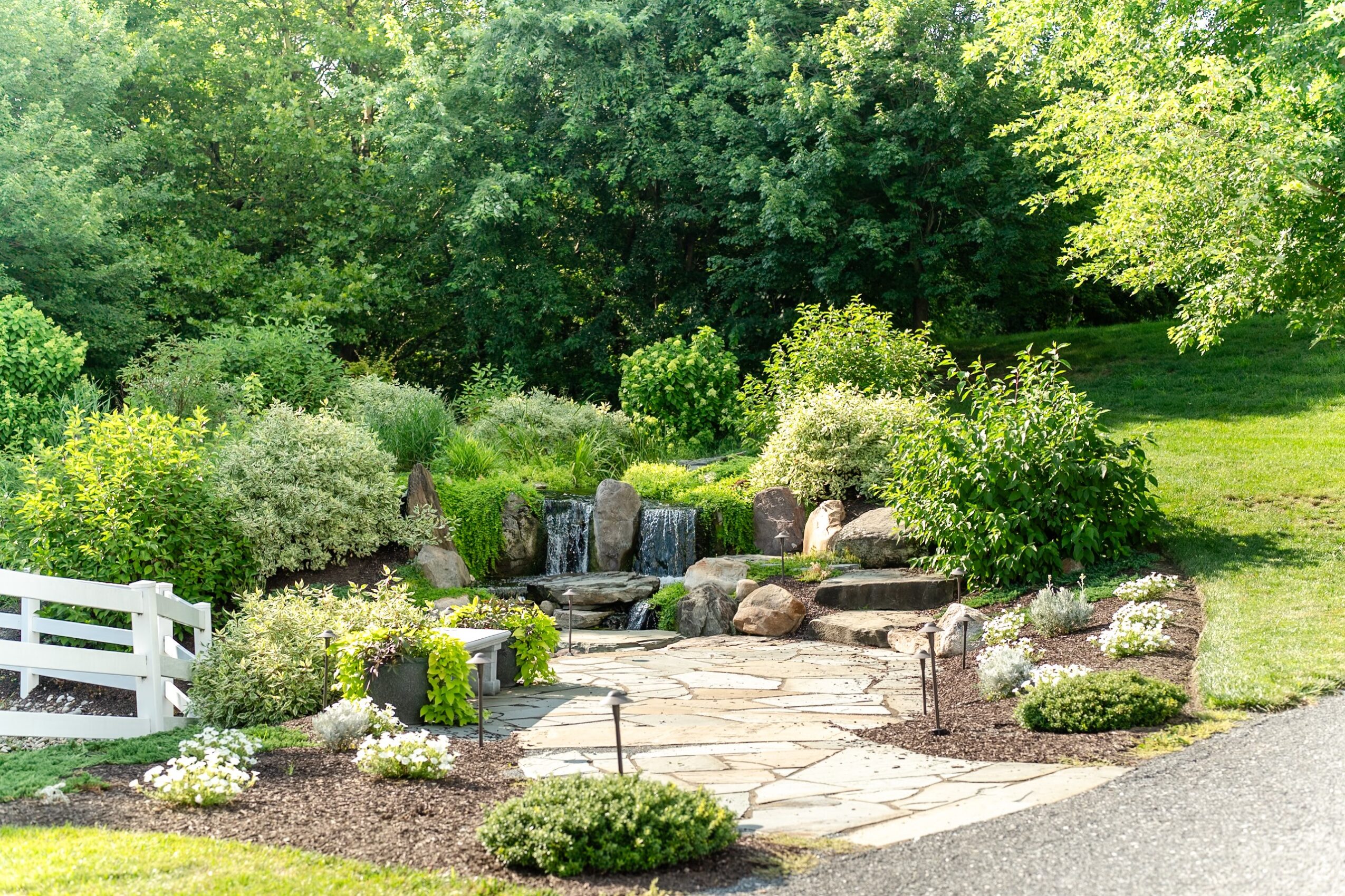 Garden with stone pathway, waterfall, lush green bushes, trees, and a white fence, under bright sunlight.