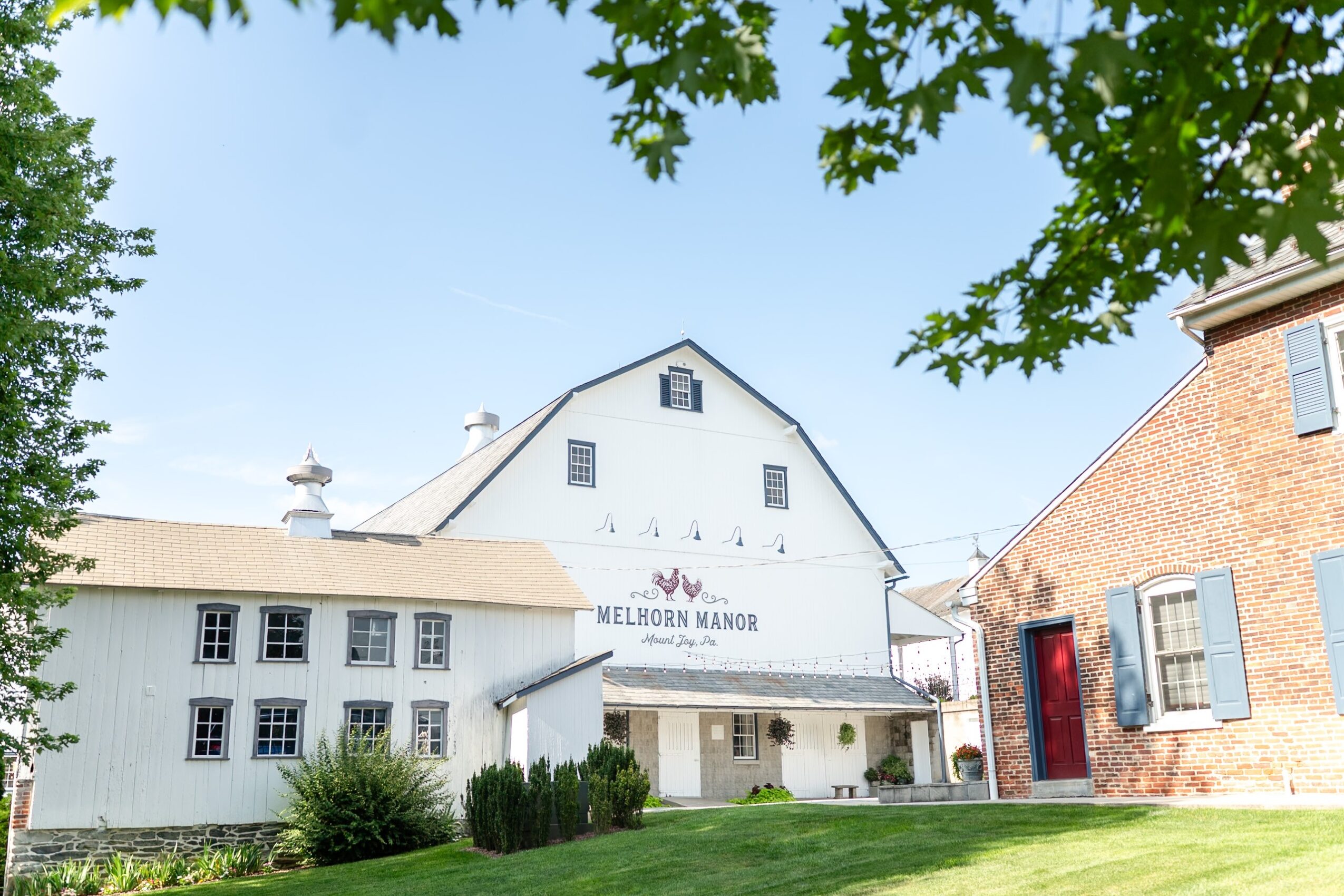 White building with sign reading Melbourn Manor, surrounded by greenery and neighboring structures, under a clear blue sky.
