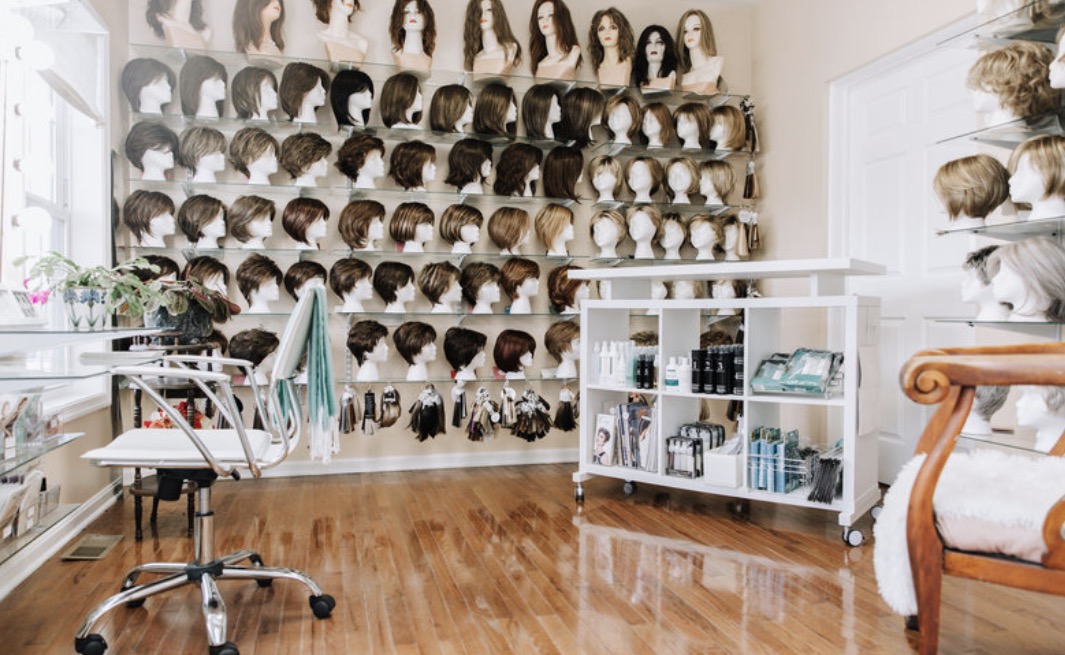 Room with shelves of mannequin heads, hair products, and a chair, with wooden flooring and a window, in a hair salon.