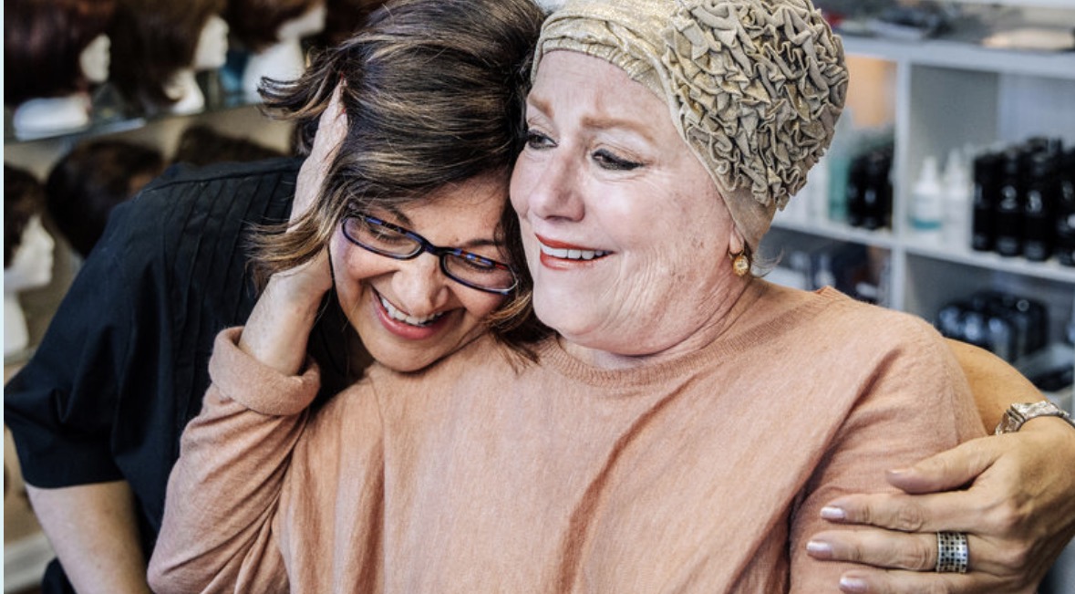 Two women hugging and smiling warmly in a store, one wearing glasses and the other with a headscarf.