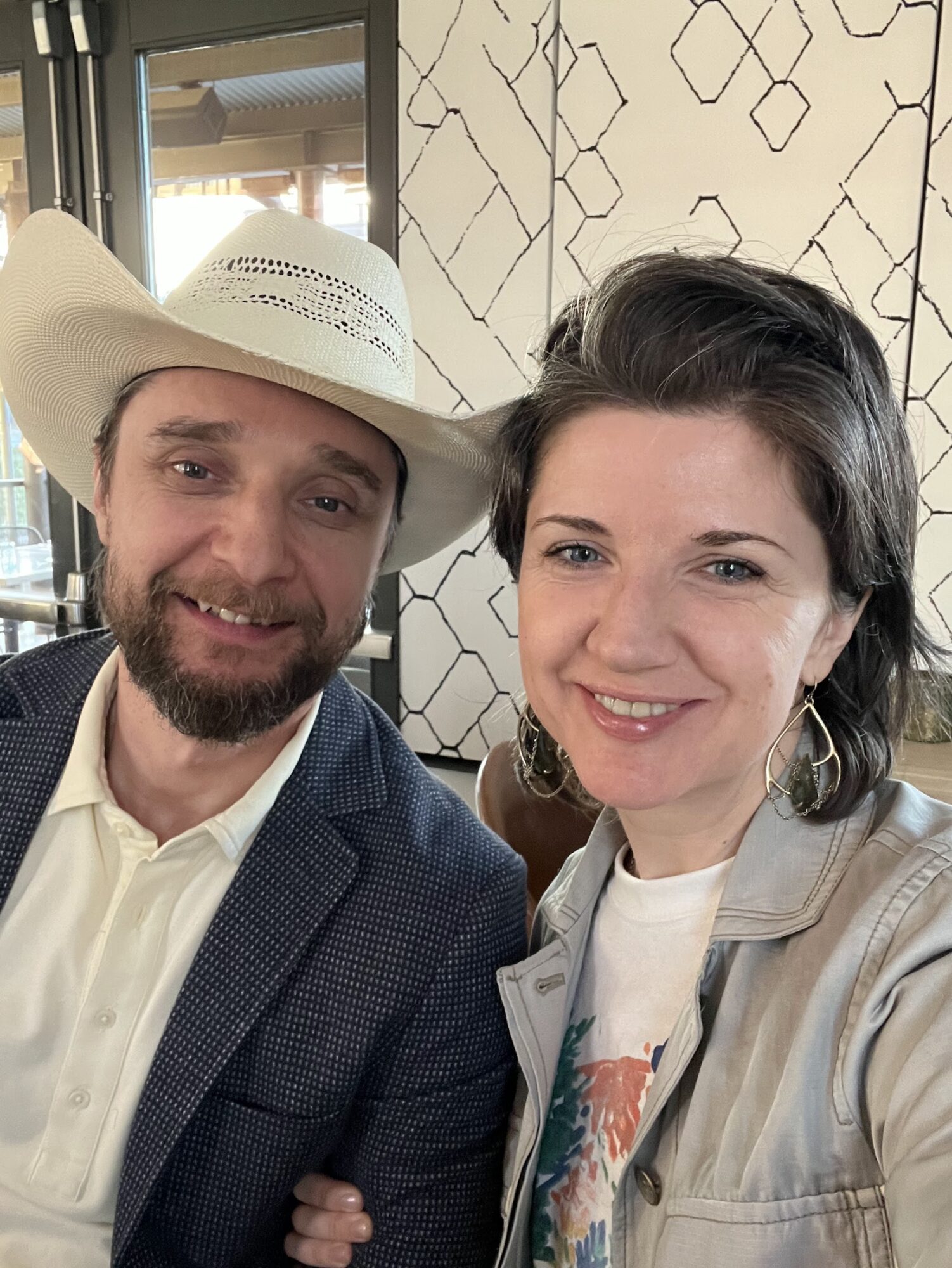 Two people smiling, one wearing a cowboy hat, in a room with geometric wall pattern.