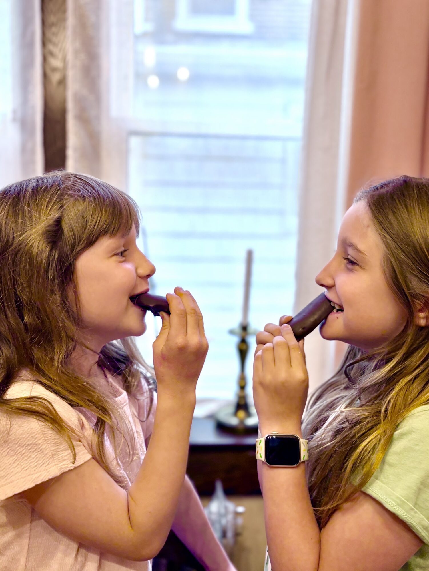 Two young girls with long hair eating ice cream cones indoors near a window, smiling at each other.