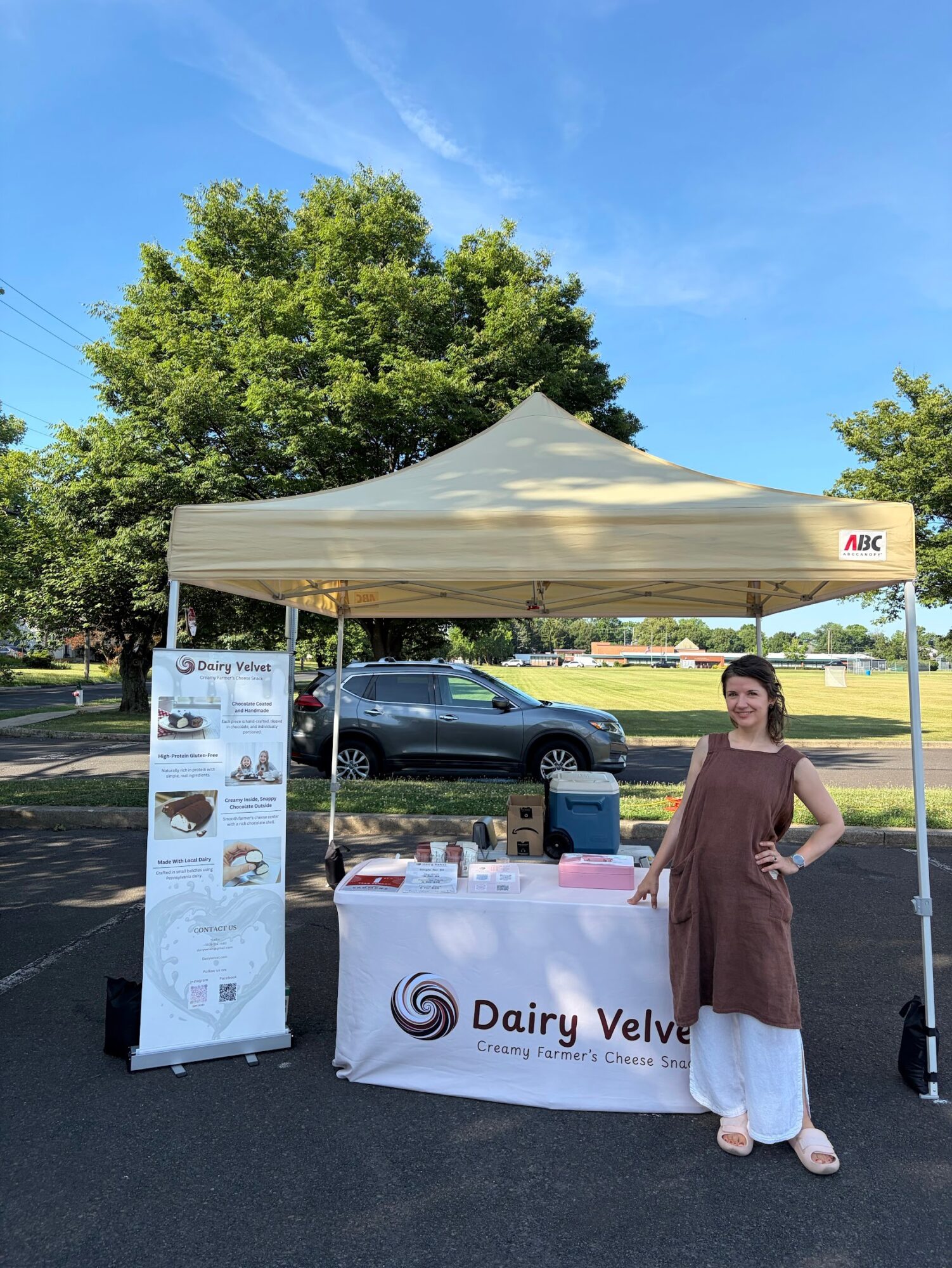 Woman standing next to a Dairy Valley booth under a white canopy, with a large tree and parked car in the background.