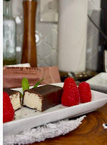 Chocolate-covered dessert with raspberries on a white plate, on a wooden table, with kitchen items in background.