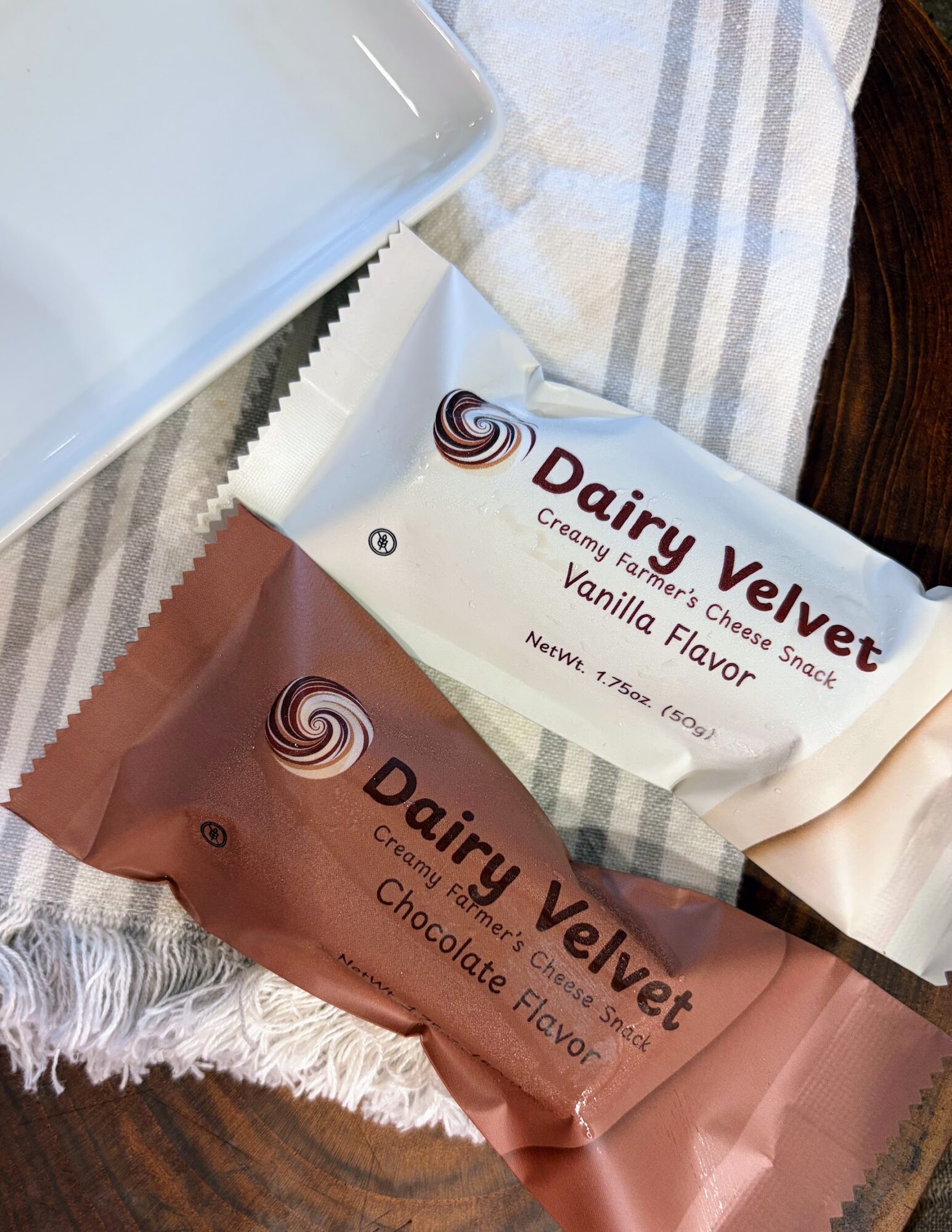 Two Dairy Velvet snack bars, one vanilla flavor and one chocolate flavor, on a cloth surface with a plastic container nearby.