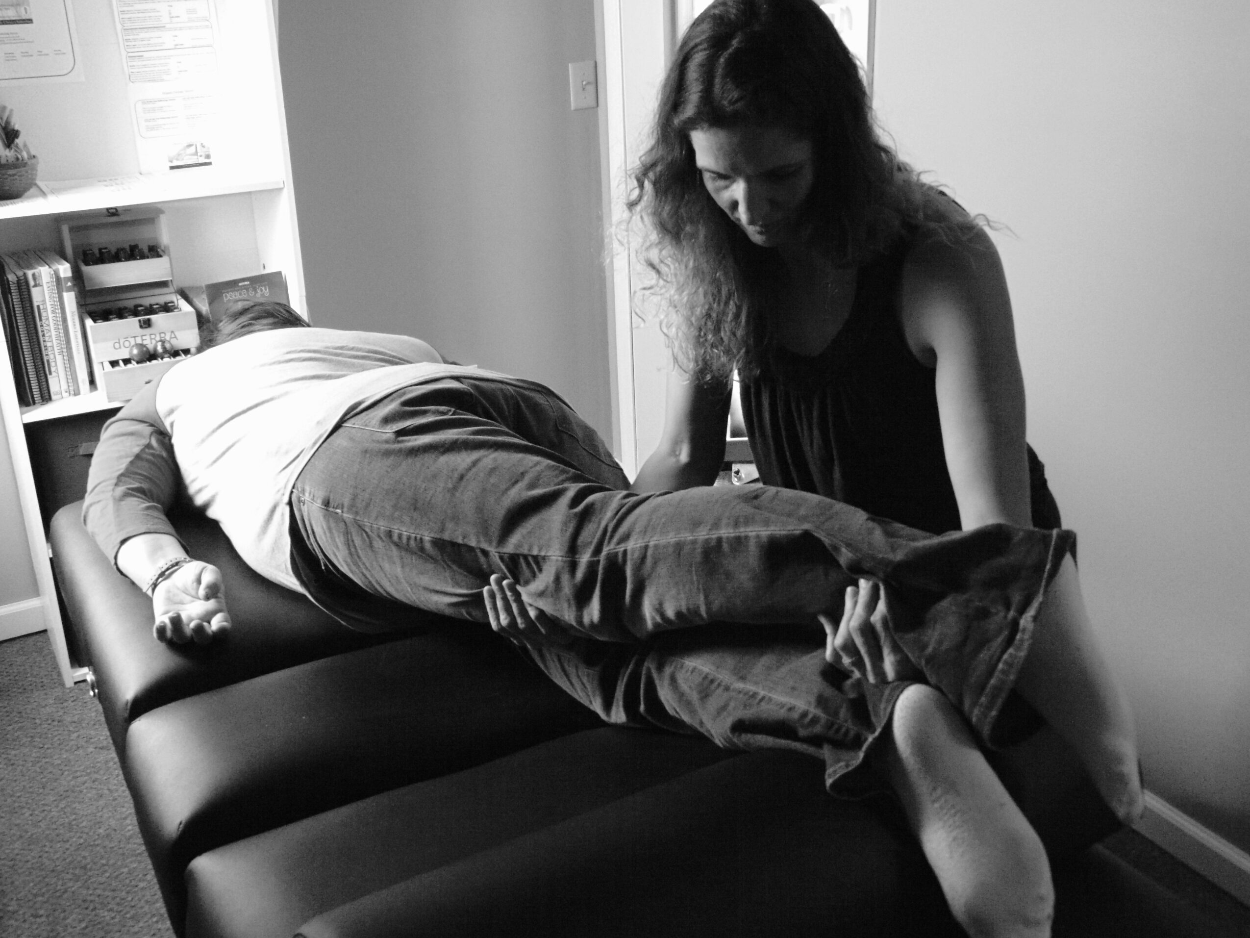 Person lying on a table with another person attending to their foot, in a room with shelves and books.