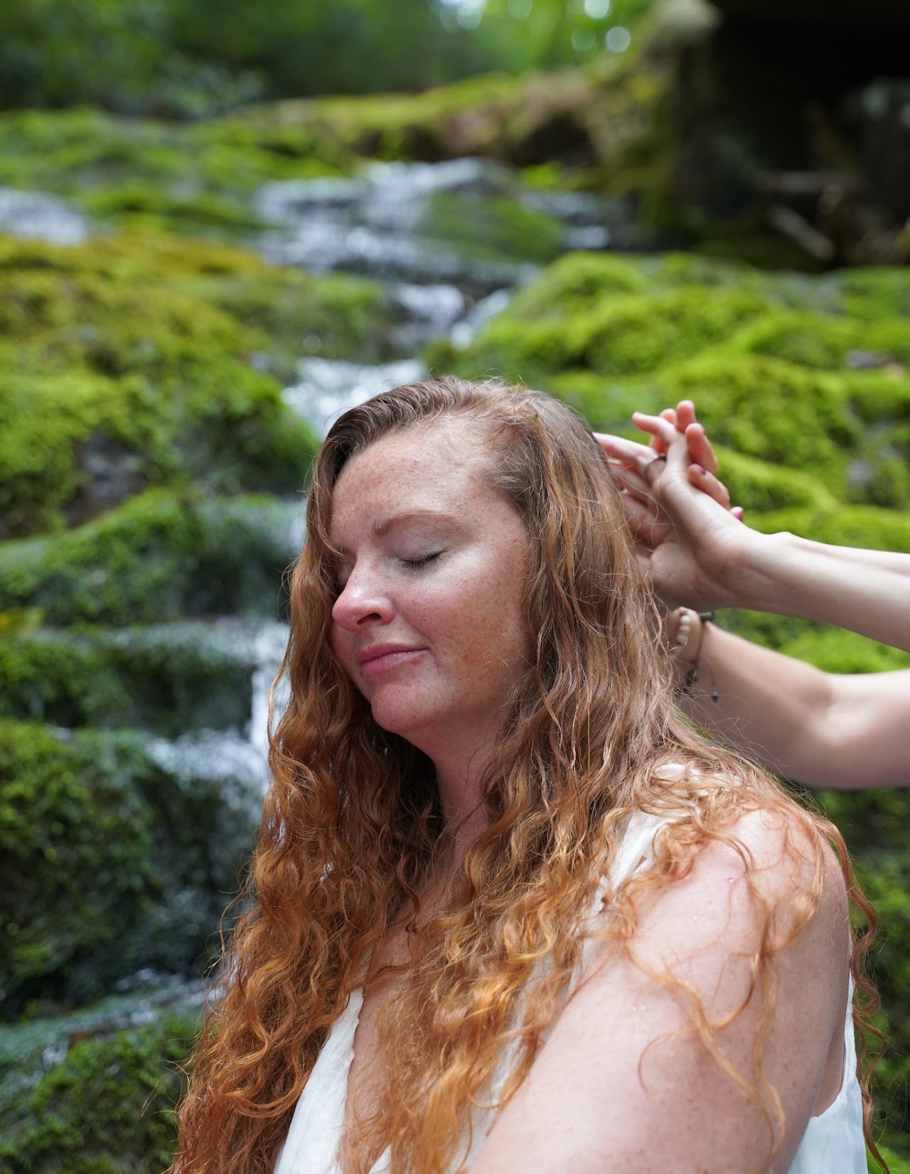 Young woman with long, curly hair relaxing outdoors near a moss-covered stream, eyes closed, peaceful expression.