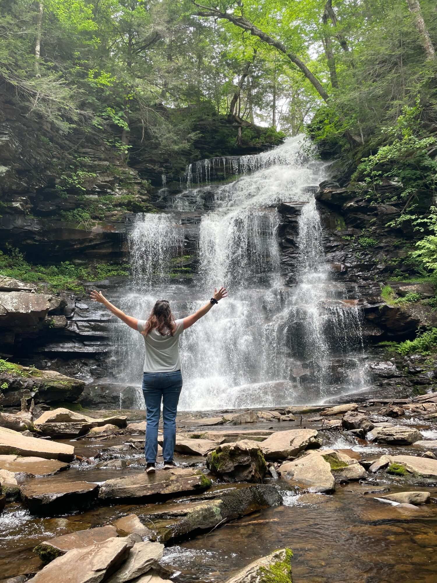 Person standing on rocks near waterfall with arms outstretched, surrounded by trees and water.