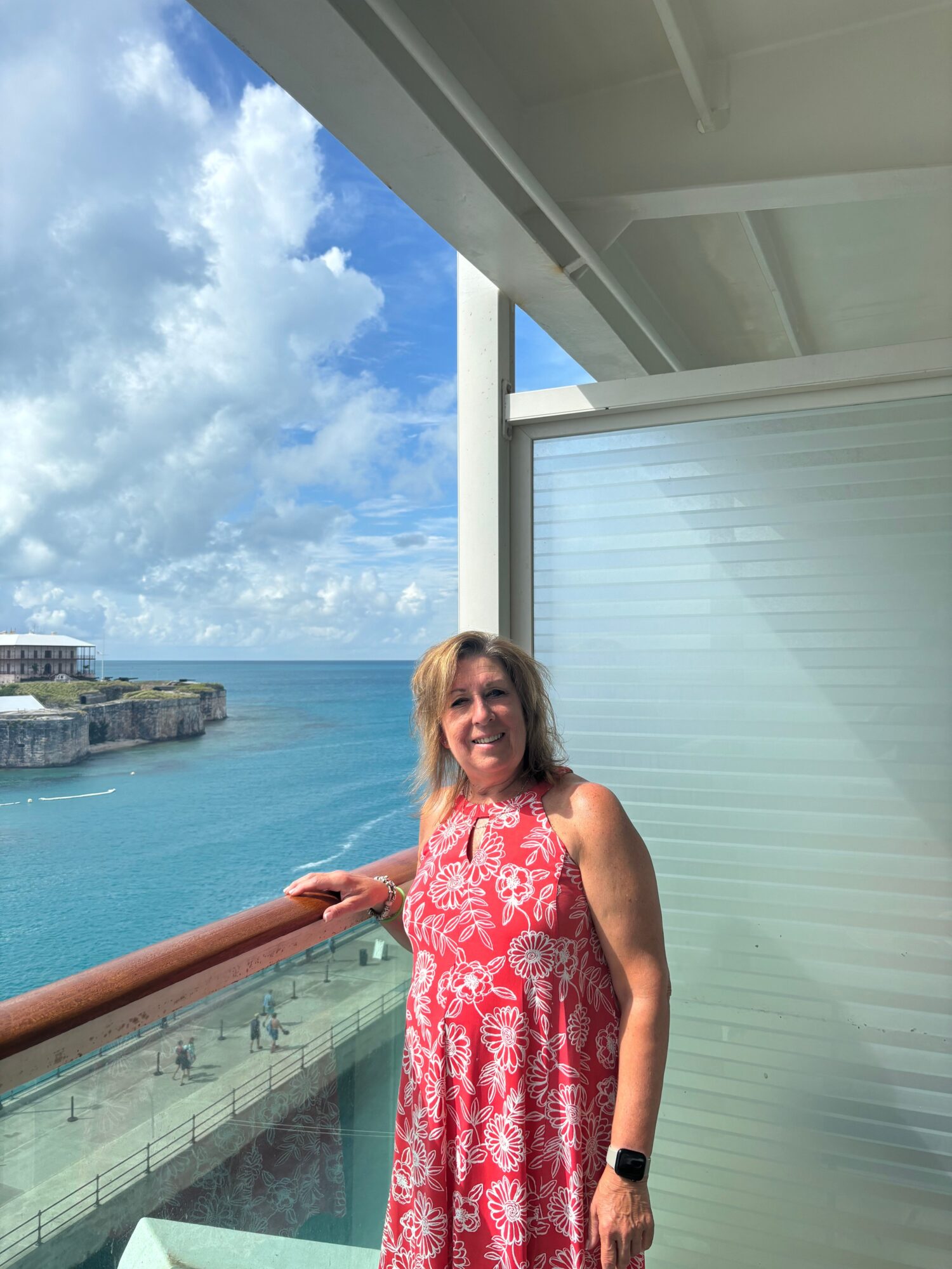 Woman in red dress standing on balcony with ocean and sky in background.