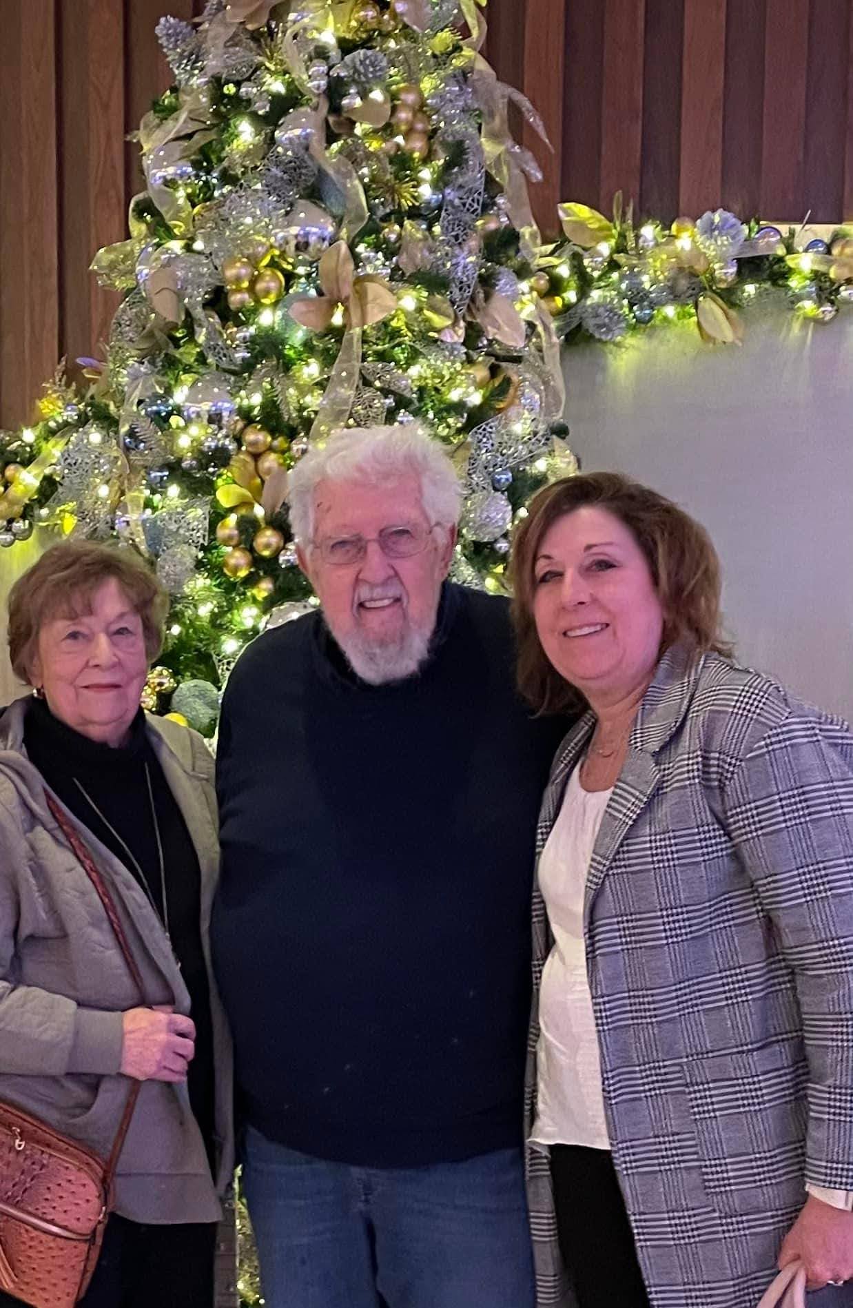 Three people standing in front of a decorated Christmas tree with lights and ornaments, smiling at the camera.