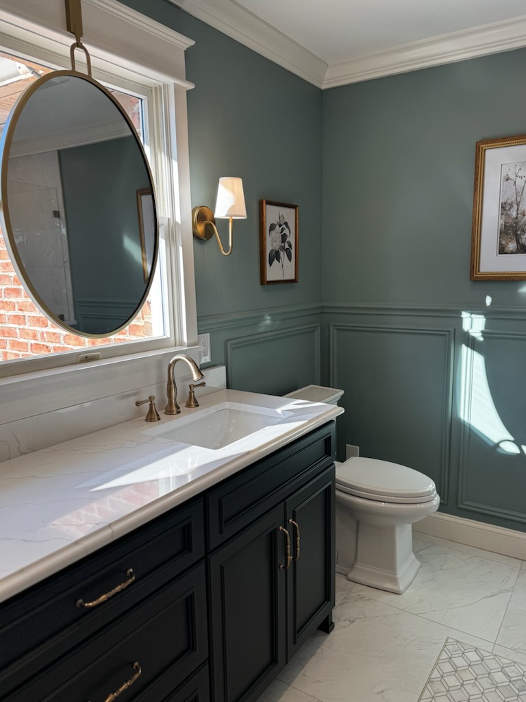 Bathroom with dark vanity, white countertop, oval mirror, green walls, framed artwork, and a toilet, illuminated by natural light.