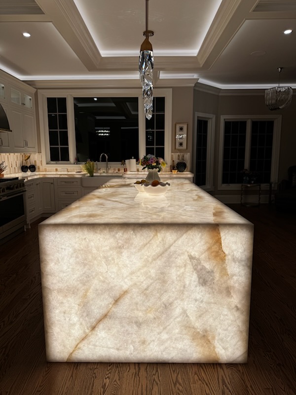 Illuminated kitchen island with a marble surface in a modern, well-lit room, with windows and ceiling lights visible.