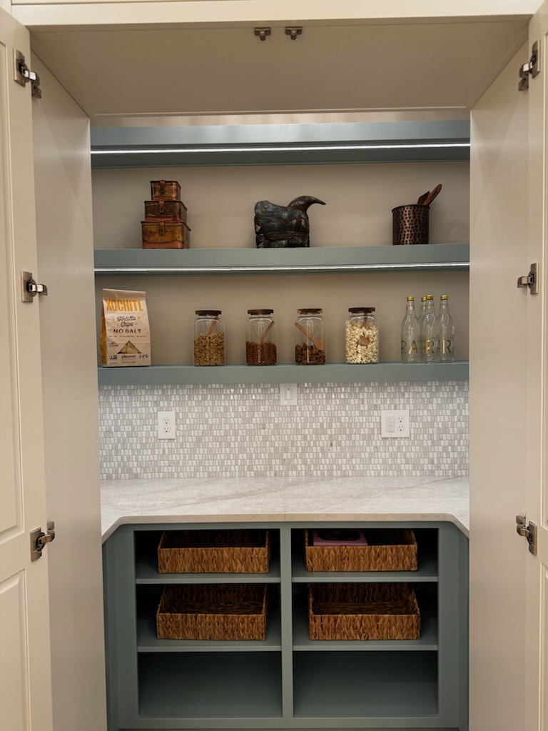 Open cabinet with shelves holding decorative items, jars, and baskets, with a tiled backsplash and storage compartments below.