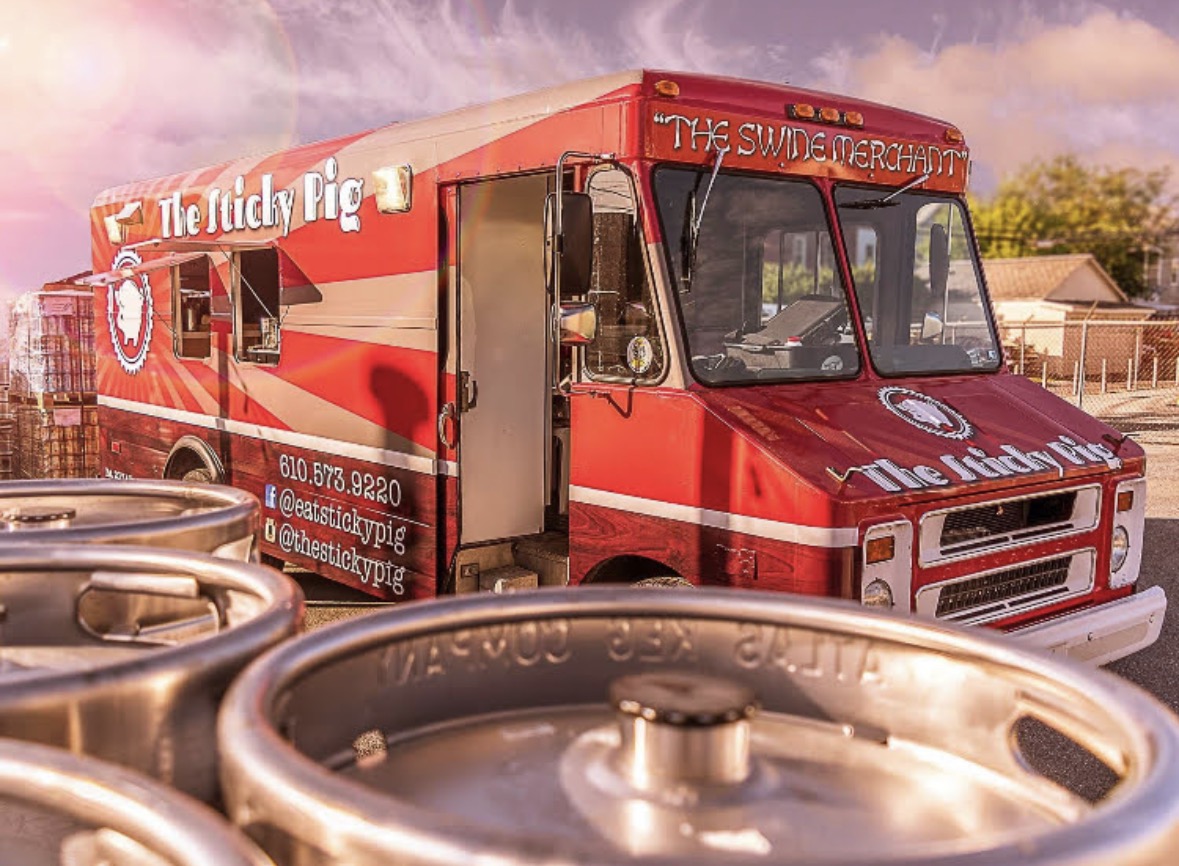 Red food truck with white and black text, parked outdoors with barrels in foreground, sky and trees in background.