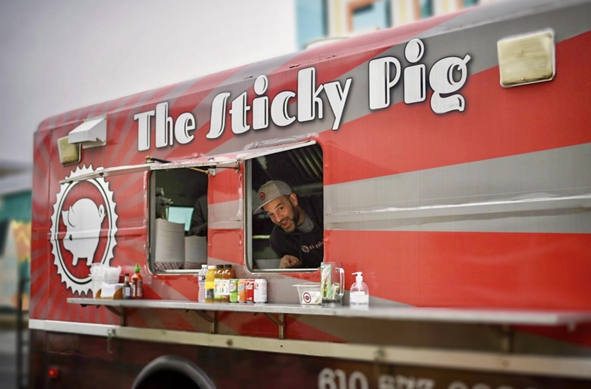 Person smiling at a food truck named The Sticky Pig with red and gray exterior, serving food through a window.