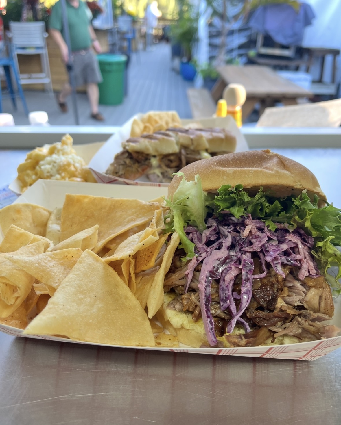 Two sandwiches with shredded meat, lettuce, and purple cabbage, served with tortilla chips on a tray, outdoor setting.