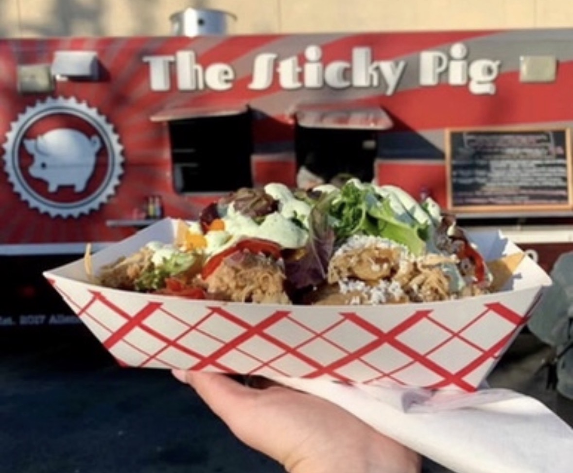 Hand holding a food tray with tacos in front of a food truck named The Sticky Pig.