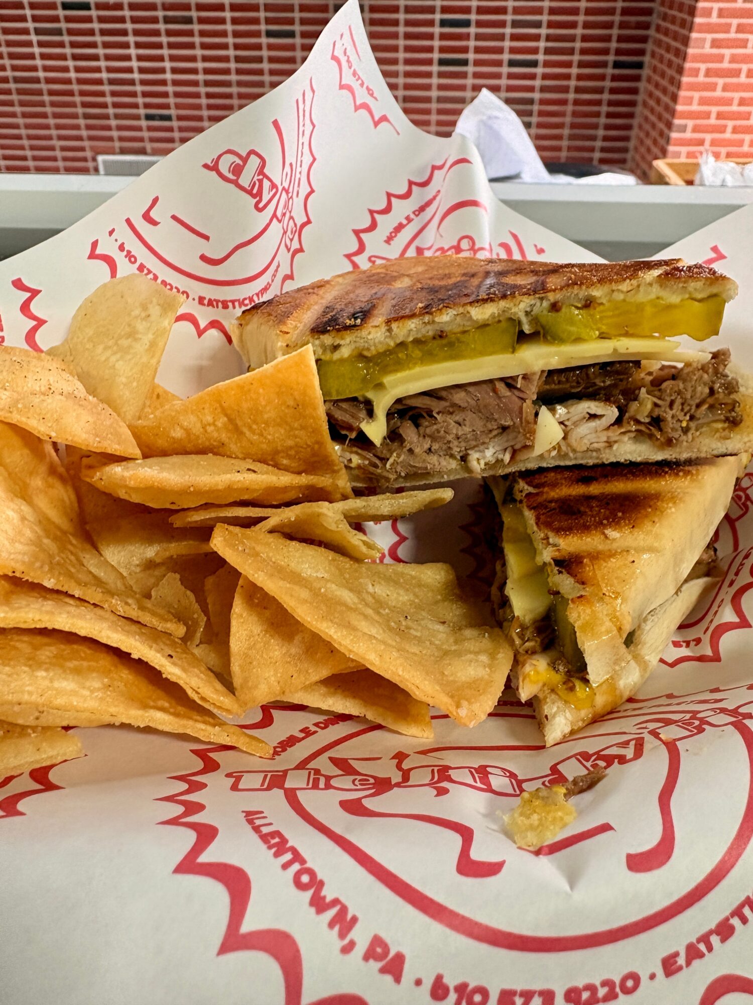 Half-eaten cheesesteak sandwich with chips on red and white paper tray, brick wall background.