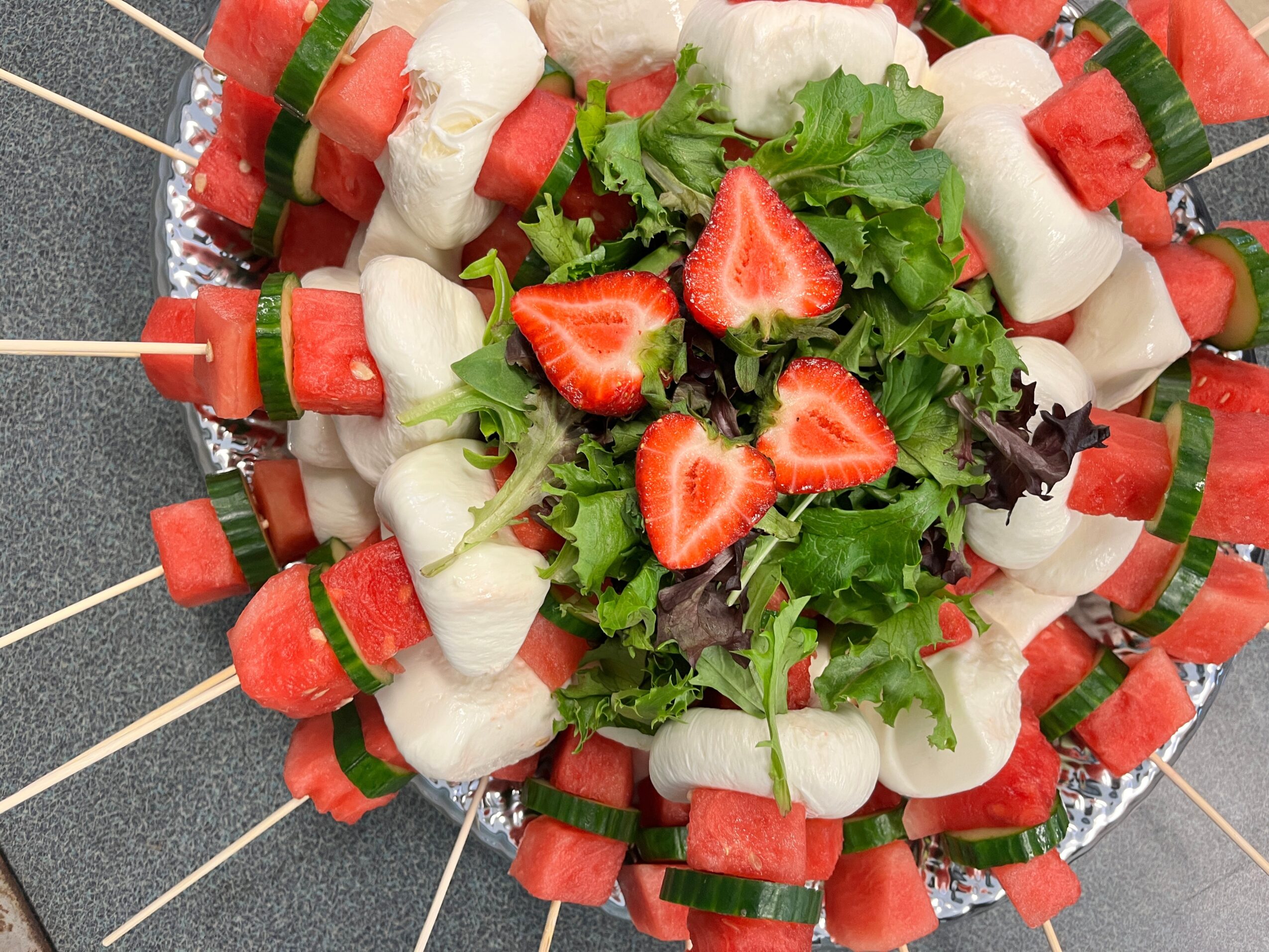 Skewers with watermelon, melon, and cucumber slices arranged around a leafy salad with strawberries in the center.