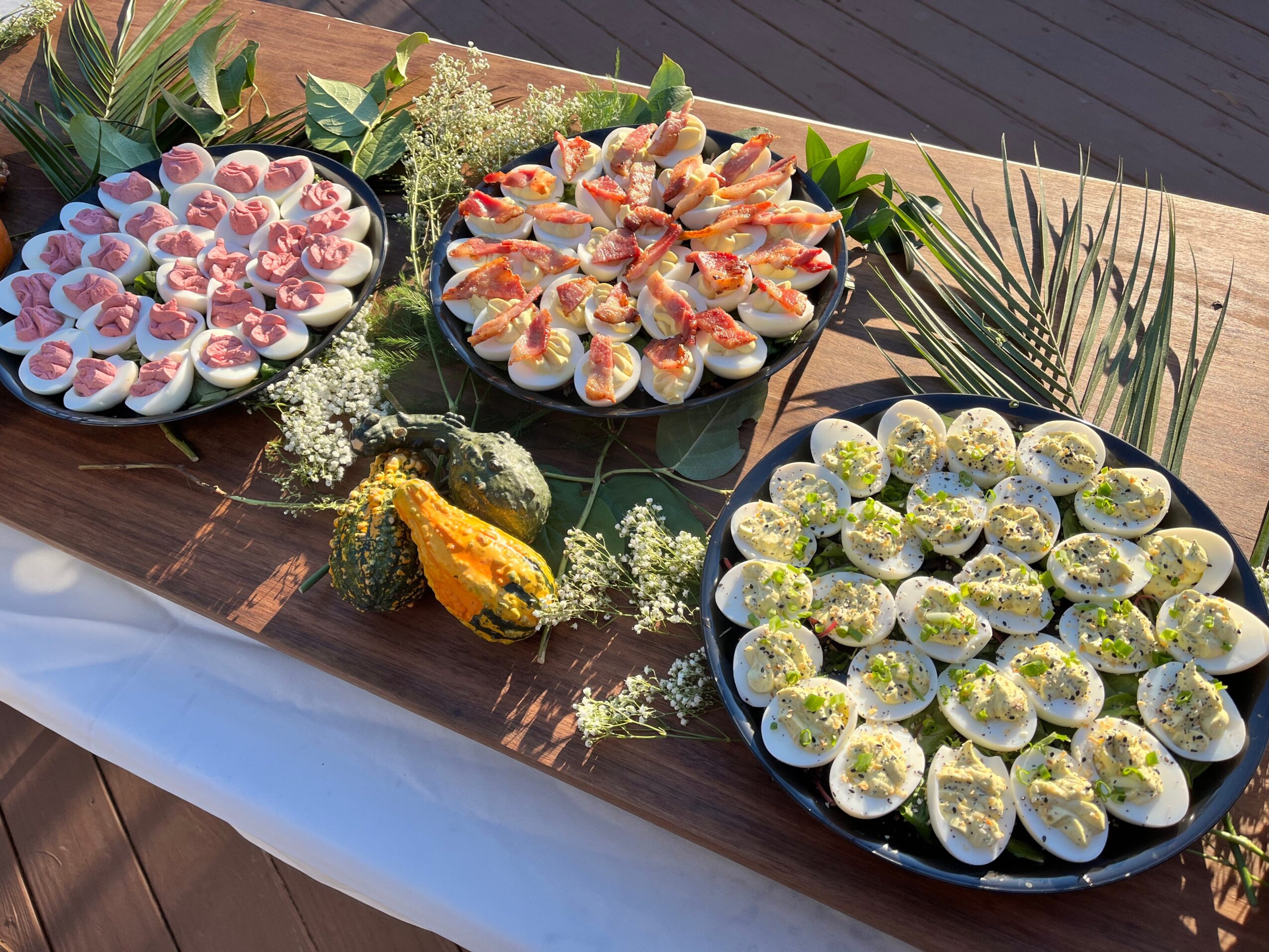 Assorted appetizers on three plates with flowers and greenery on a wooden table, outdoors.