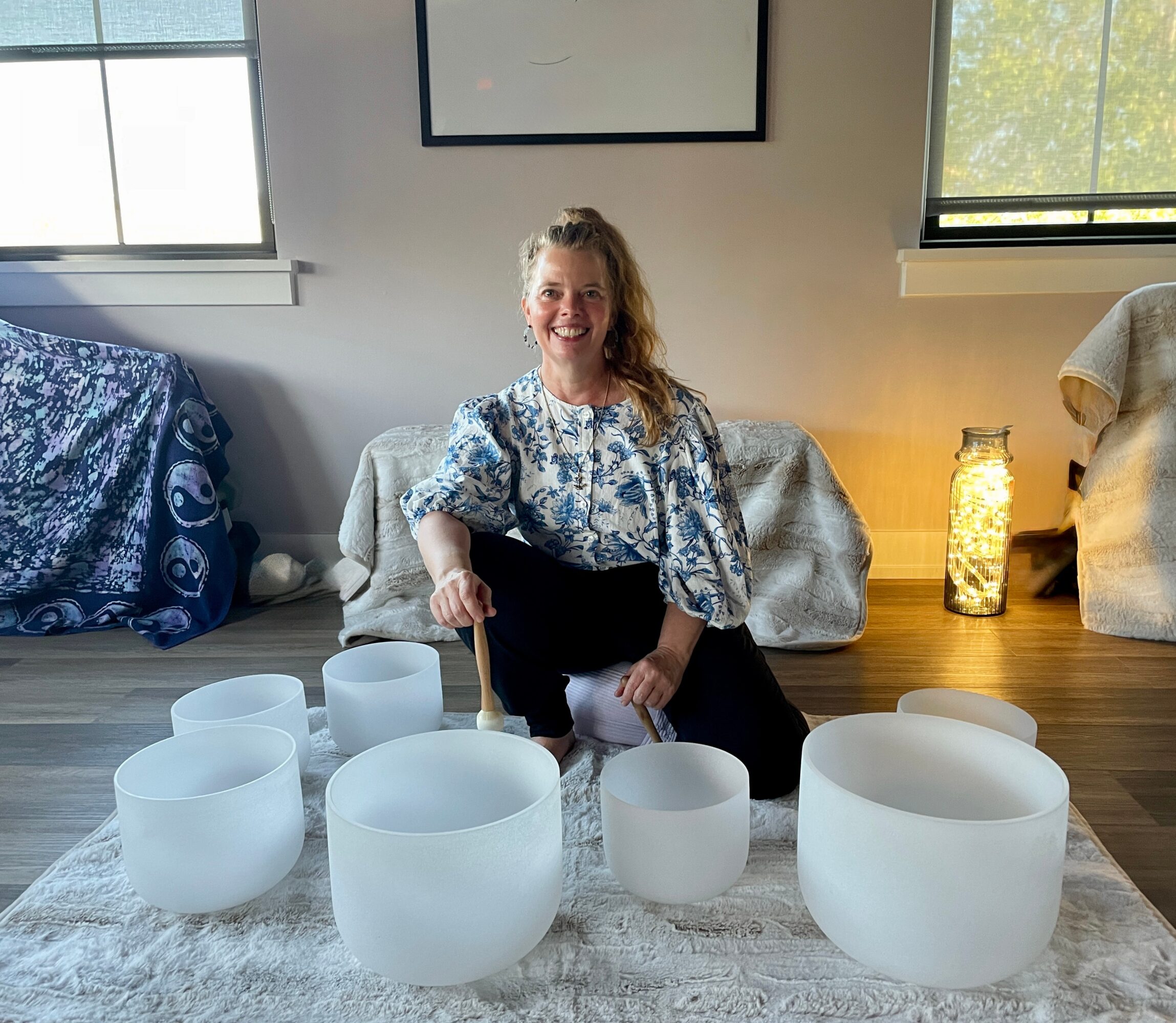 Woman sitting on the floor smiling, surrounded by white crystal singing bowls, in a room with windows and decorative lamps.