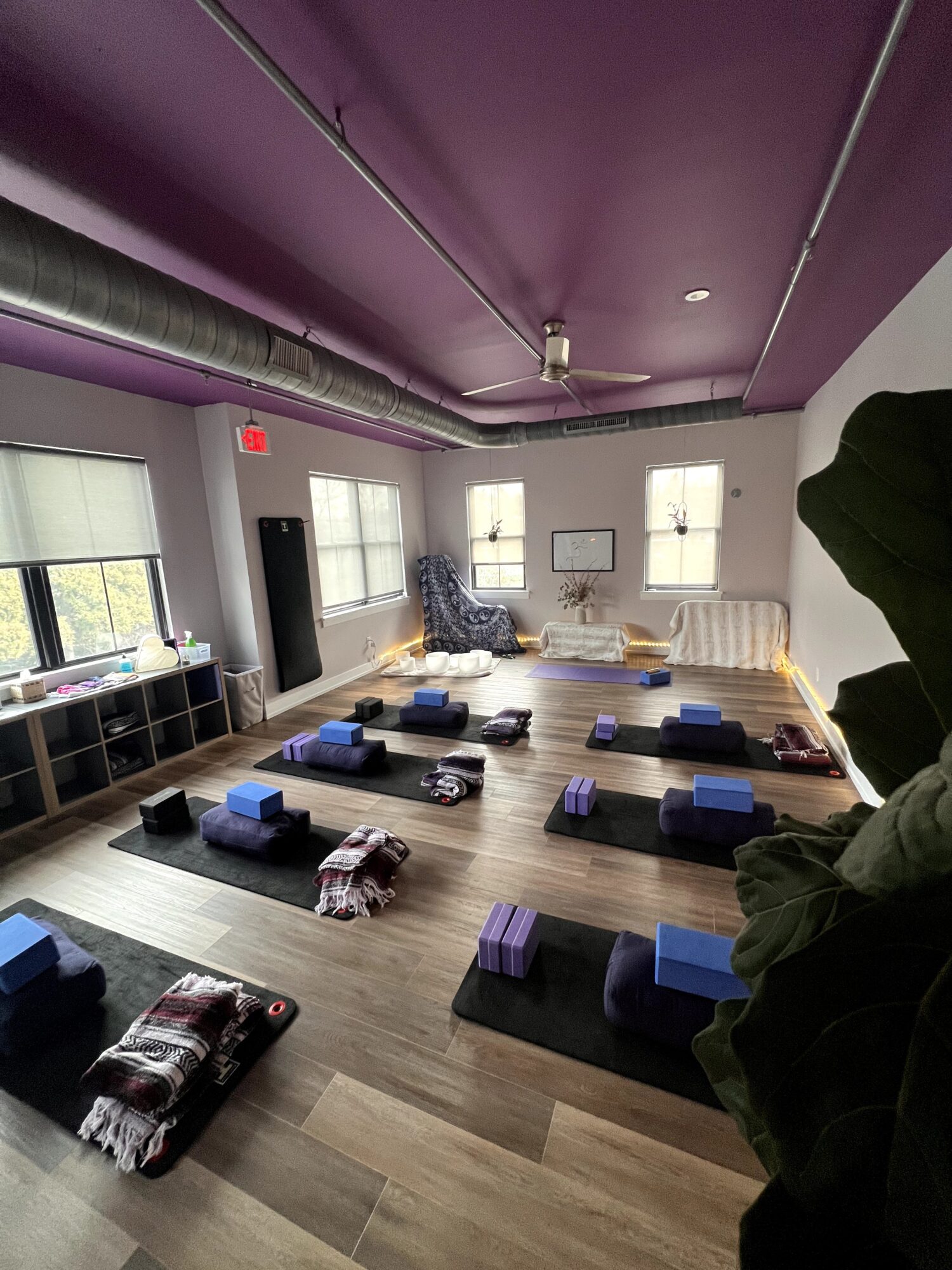 Yoga studio with mats, blocks, and blankets arranged on wooden floor, purple ceiling, and windows letting in natural light.