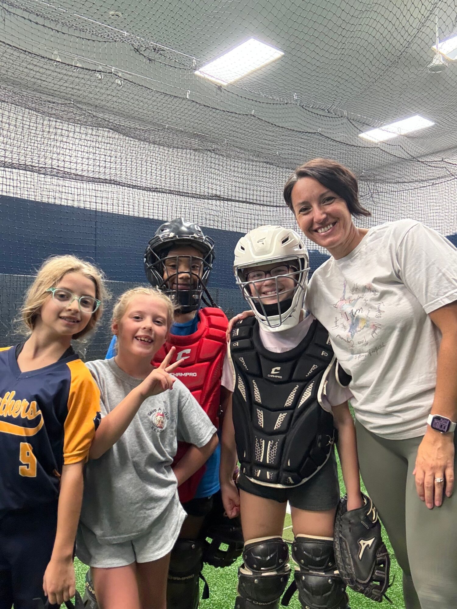 Four young women posing together indoors, one in hockey gear, others in casual clothing, smiling and making peace signs.
