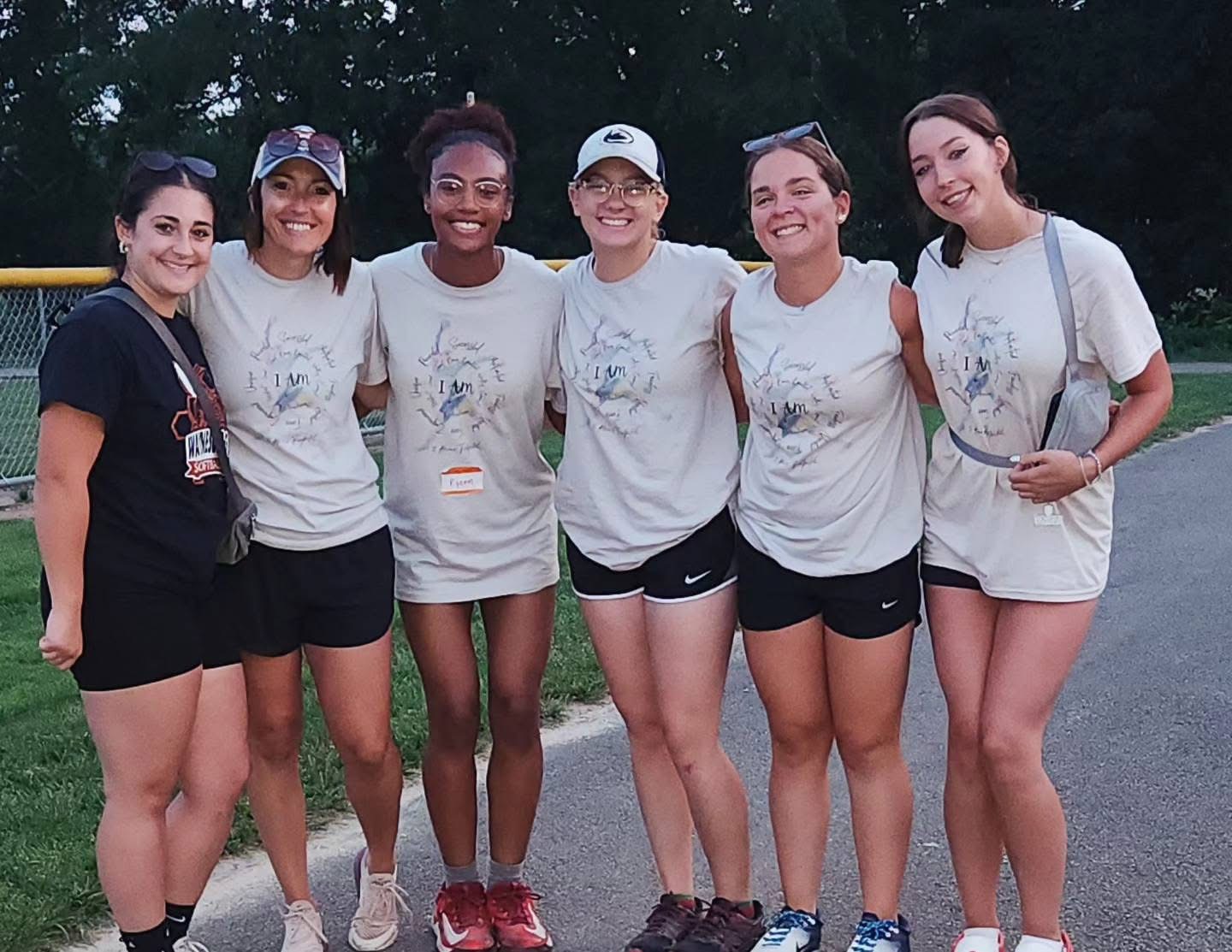 Six young women standing outdoors, smiling, wearing athletic clothing and caps, with trees in the background.