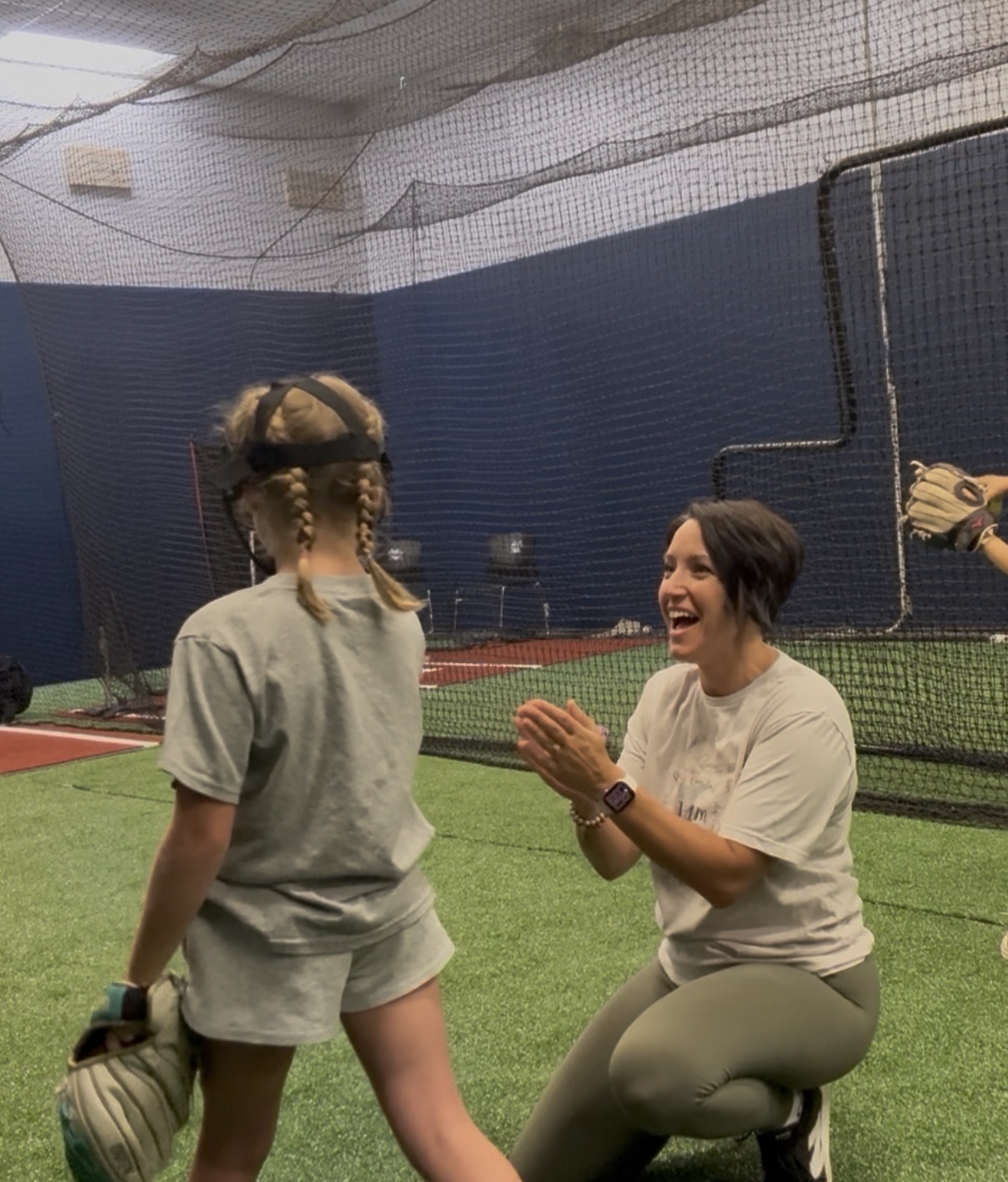 A woman kneeling on the ground claps and smiles at a girl in a sports facility with artificial turf and netting.